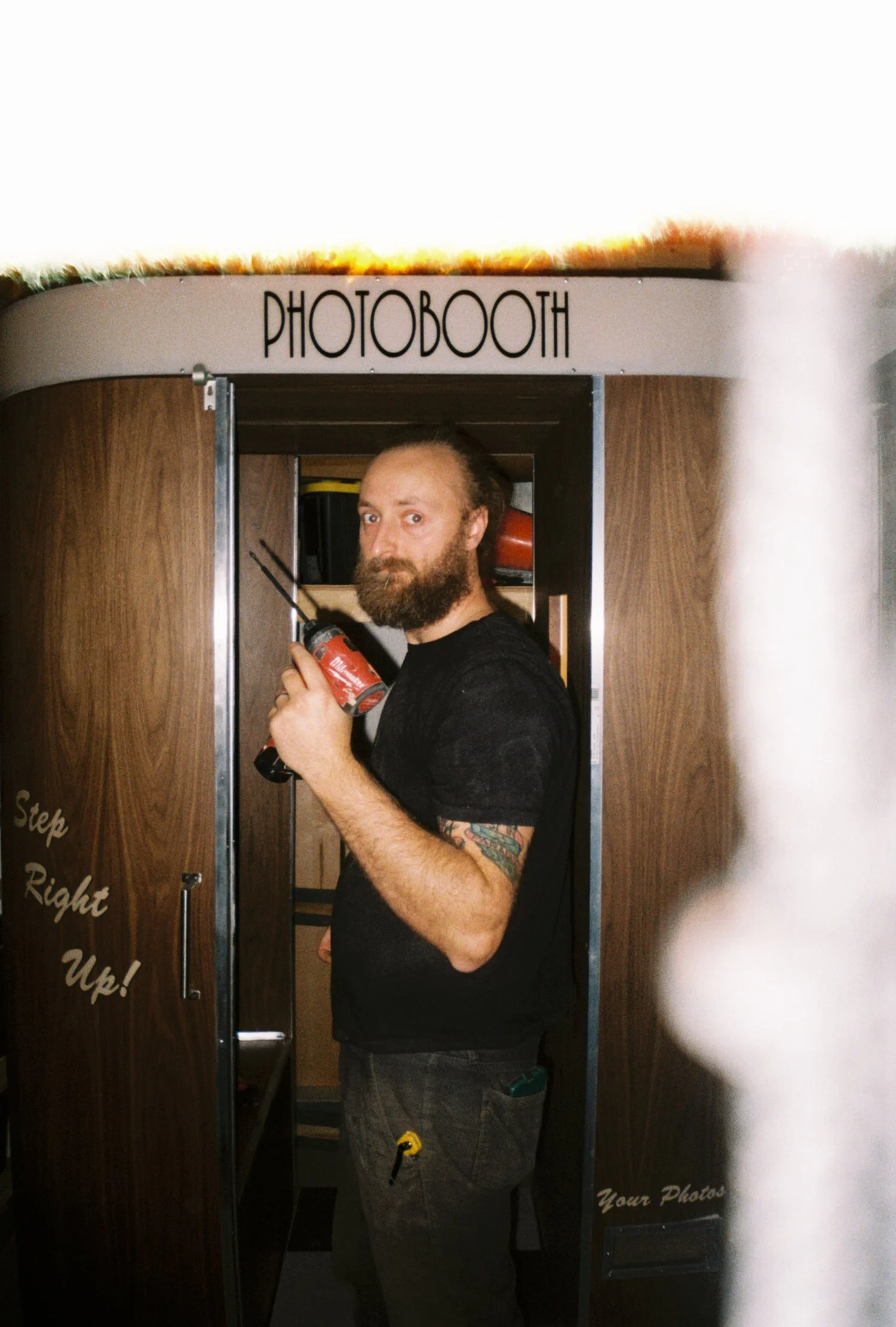 A man with a beard and a tattoo on his right arm standing inside a photo booth labeled 'PHOTOVOICE,' holding a red caulking gun, with a sign on the door that reads 'Step Right Up!'