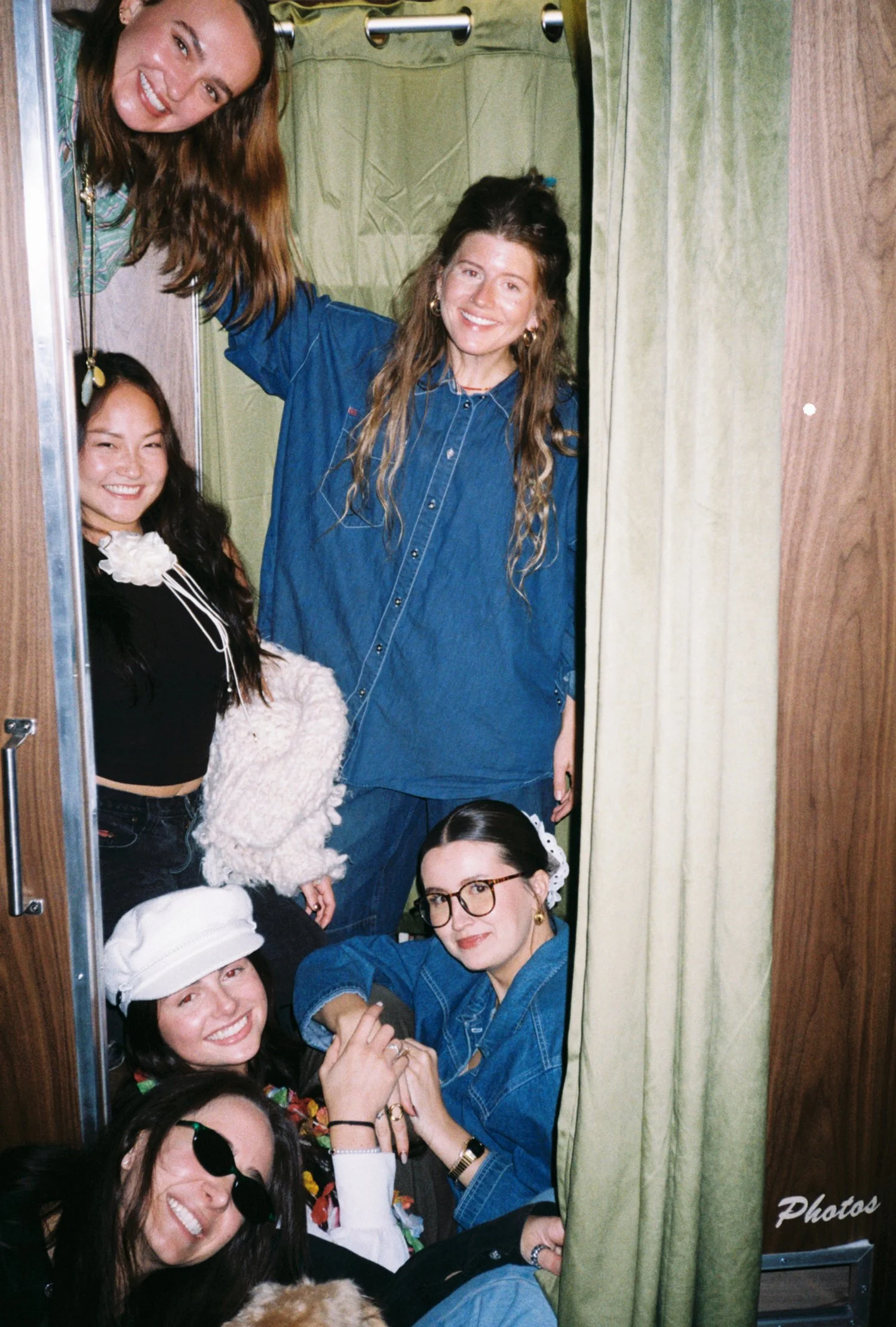 Six women in a photo booth, smiling and posing. One woman with long brown hair in a plaid shirt leaning out of the top. A woman with long dark hair wearing a black top, a fluffy white sweater, and a white hat. A woman with glasses and a denim jacket 