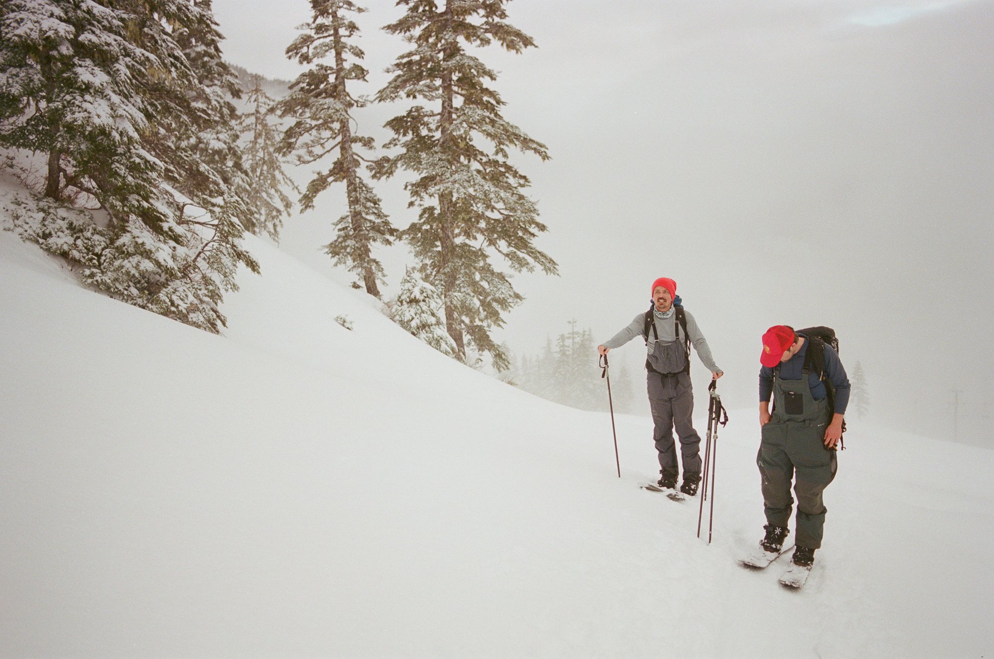Two people with red hats skiing uphill in a snowy forest.
