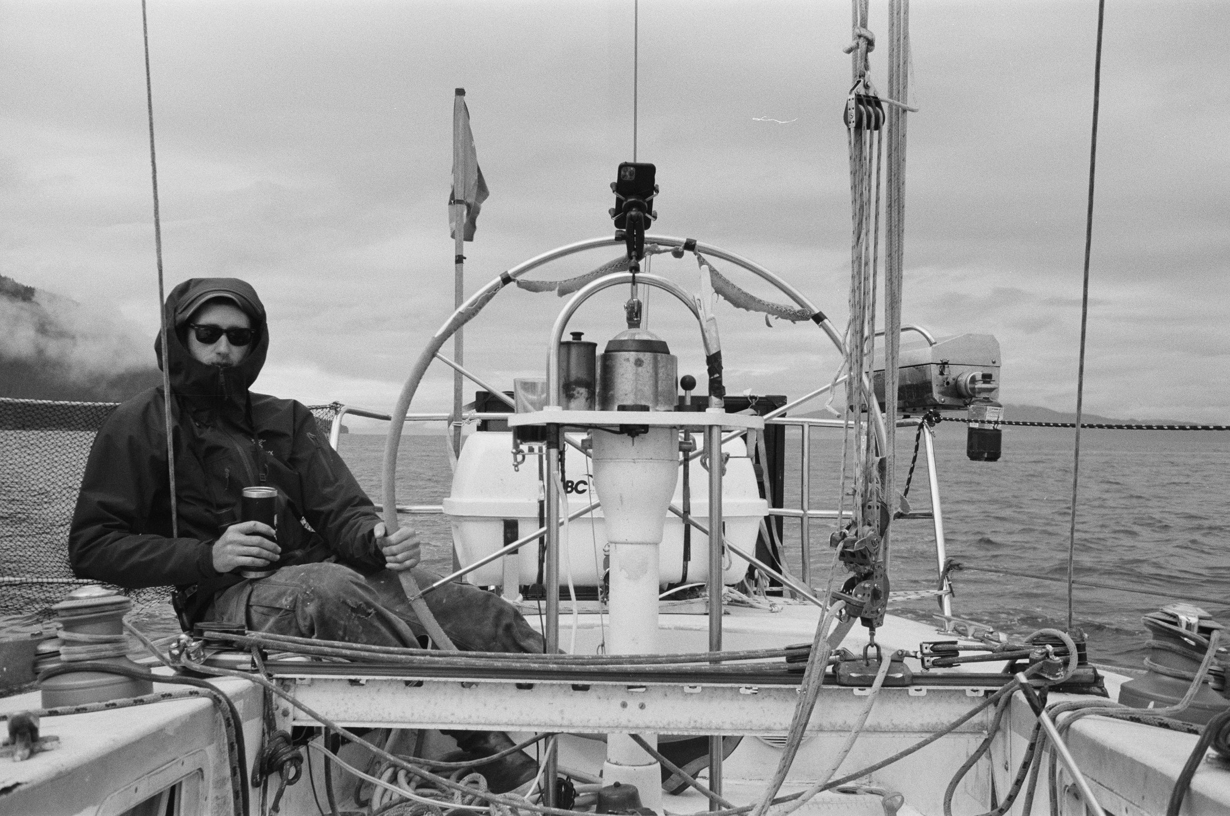 Person in hooded jacket and sunglasses holding a drink, sitting on a sailboat with a view of the ocean and cloudy sky.