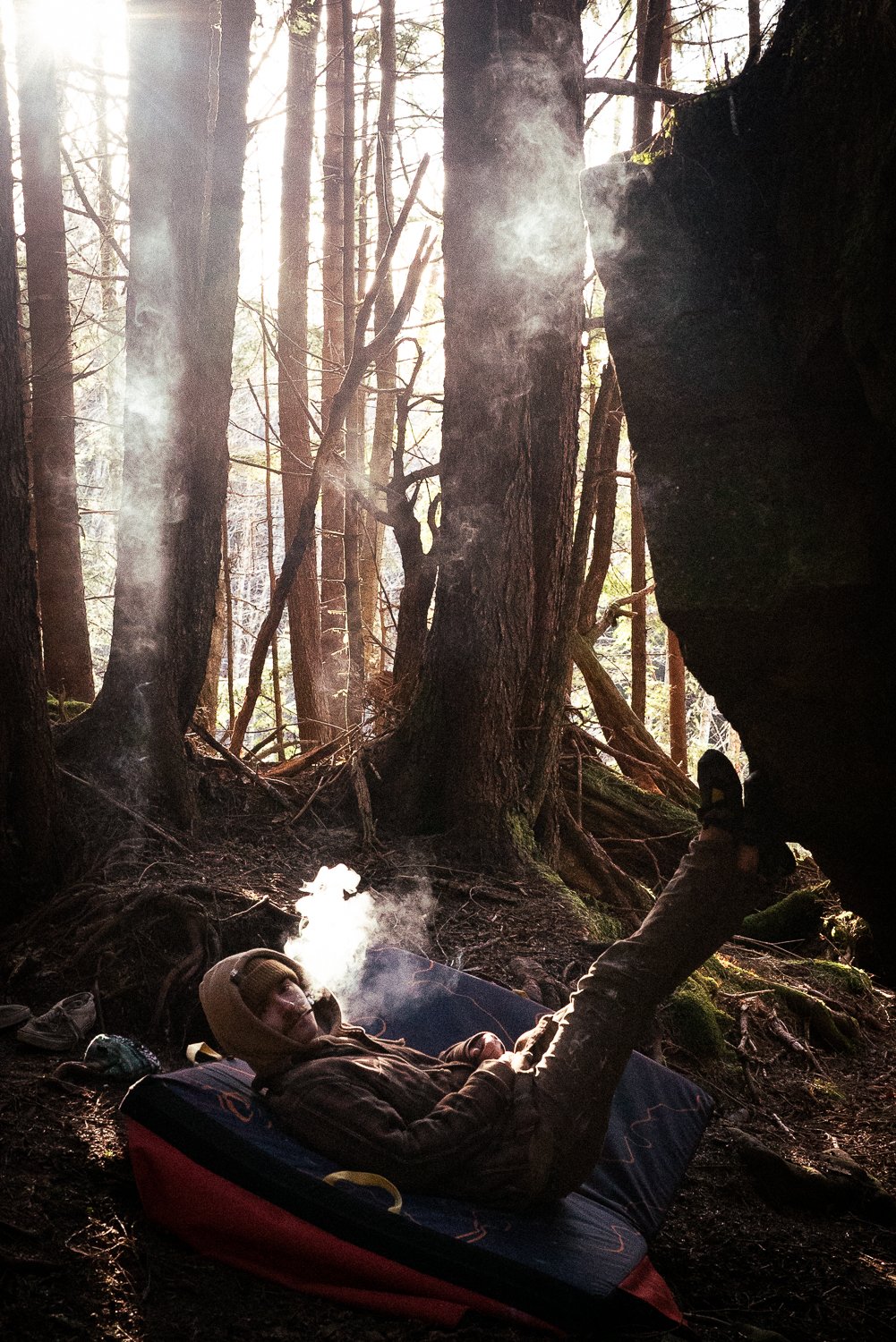 Person laying on a crash pad in the forest, exhaling smoke, surrounded by trees and sunlight.