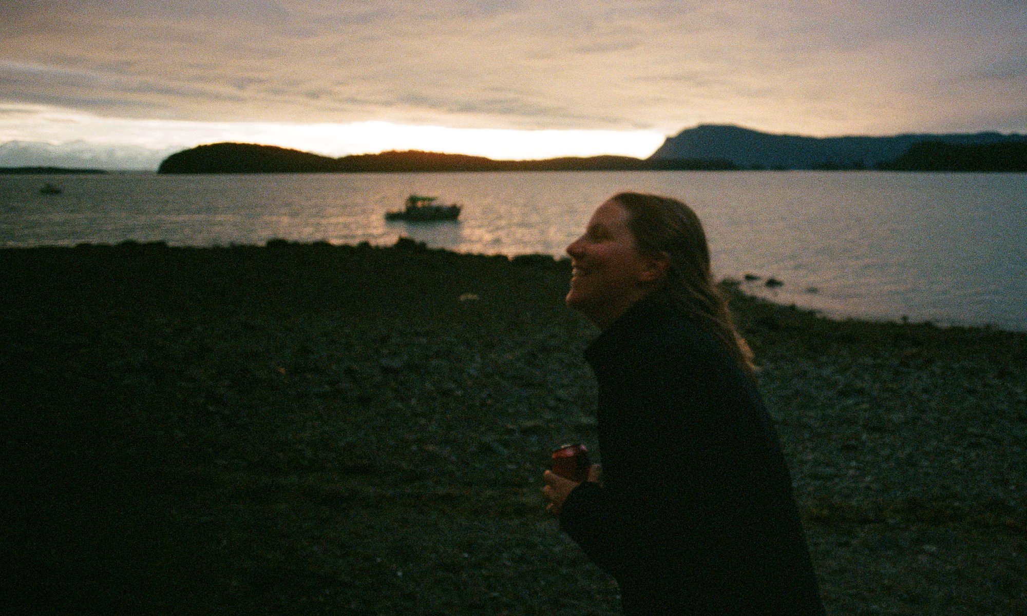 Silhouette of a person laughing by a beach, holding a drink, with a boat on water and a sunset in the background.
