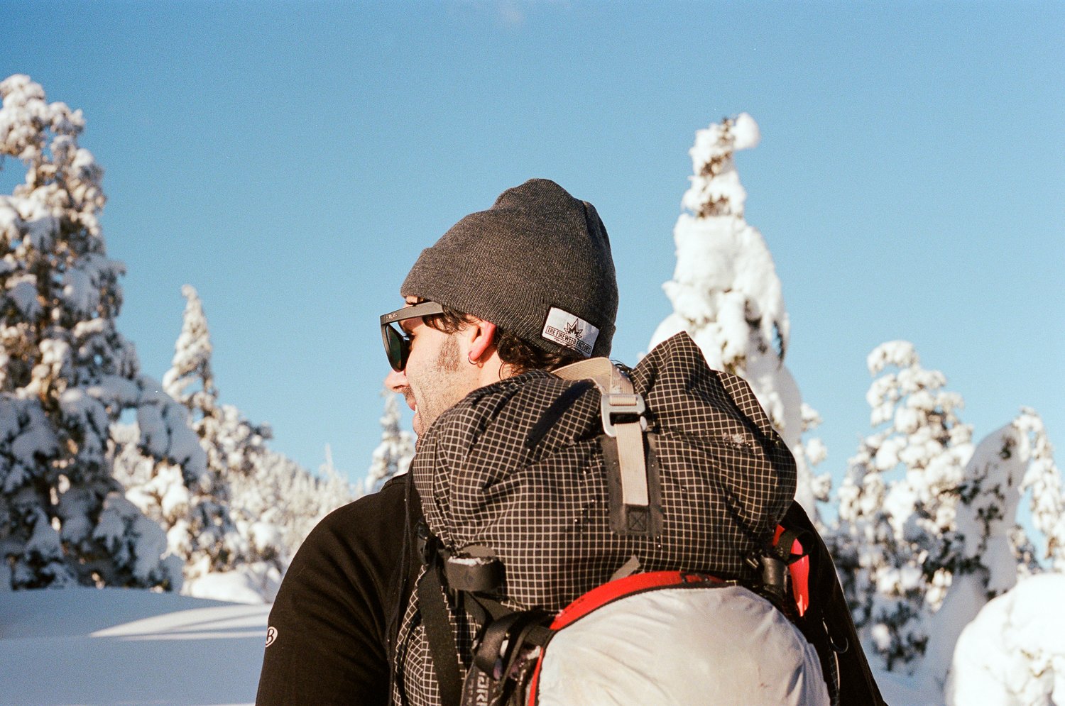 Person wearing a beanie and sunglasses carrying a backpack in a snowy landscape with snow-covered trees.