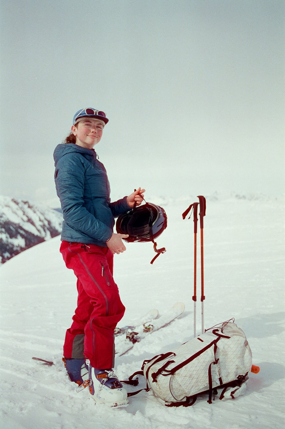 Person on snow holding a ski helmet, wearing a jacket, and standing near ski poles and backpack.