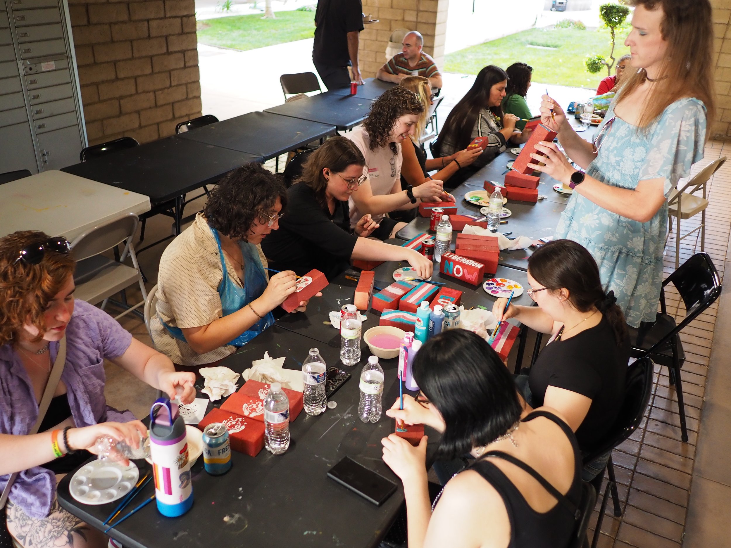 About ten folks around a long table, painting foam bricks for a pride event in 2025