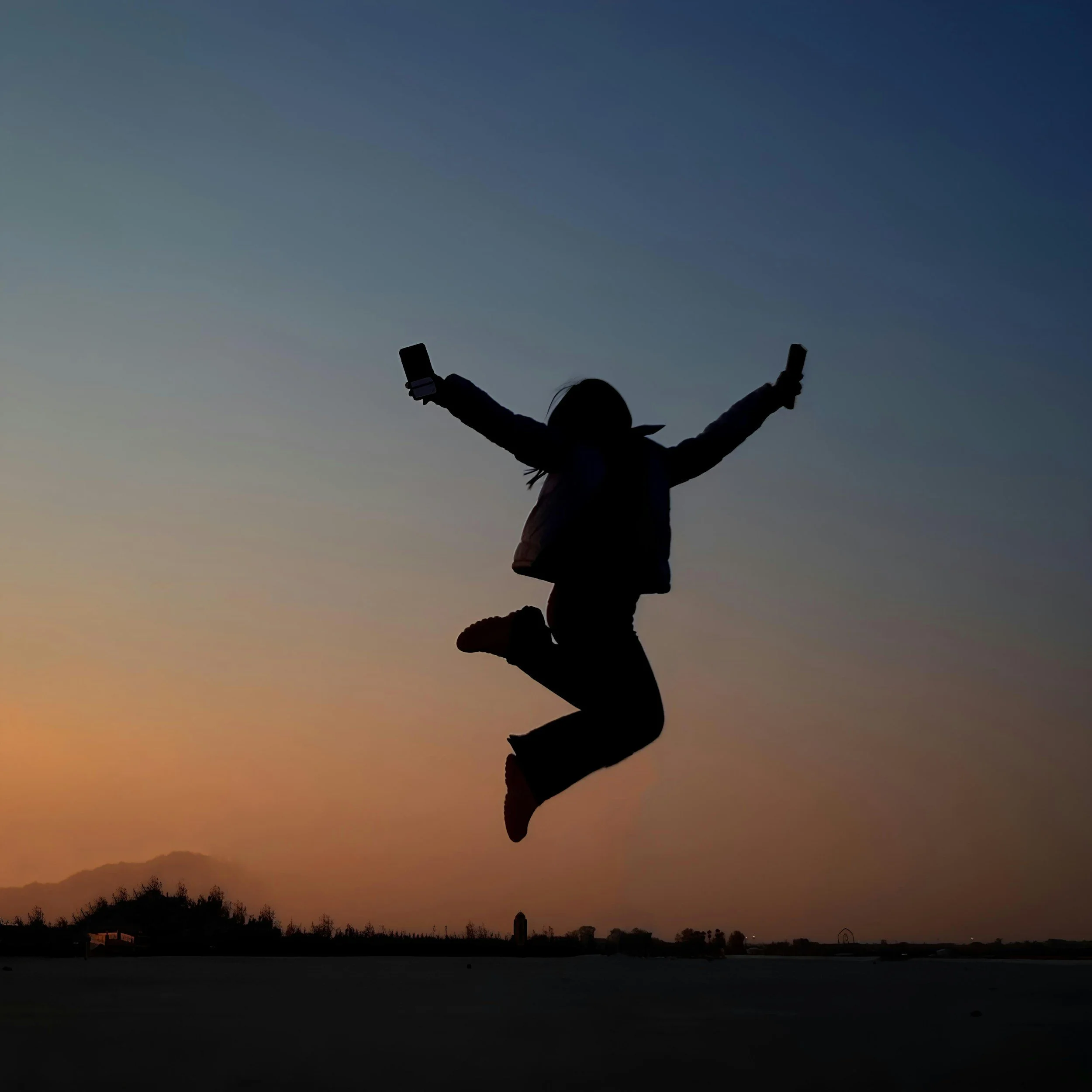 Silhouette of a person jumping with arms raised holding two phones, against a sunset sky with mountains and trees in the background.