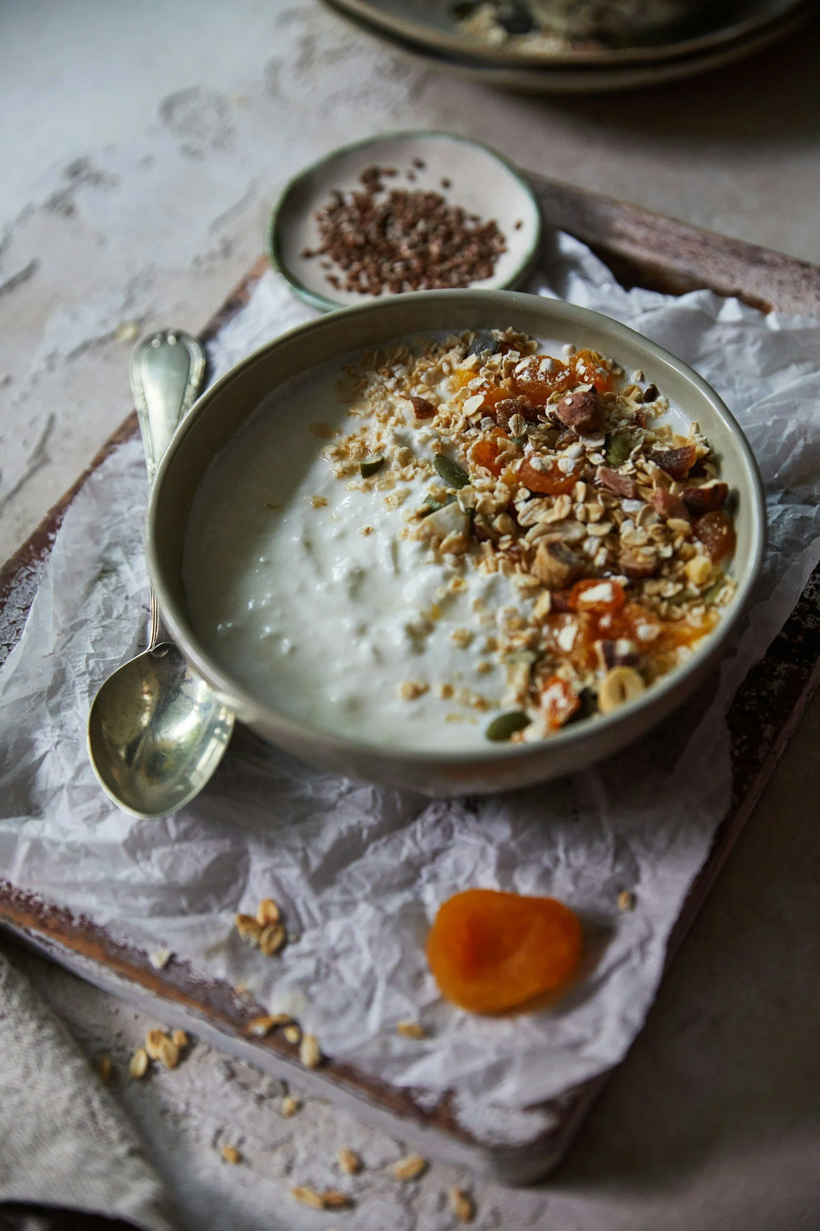 A bowl of yogurt topped with granola and mixed nuts, placed on crumpled parchment paper on a rustic wooden surface. In the background, there is a small bowl of additional granola and a long-handled spoon.