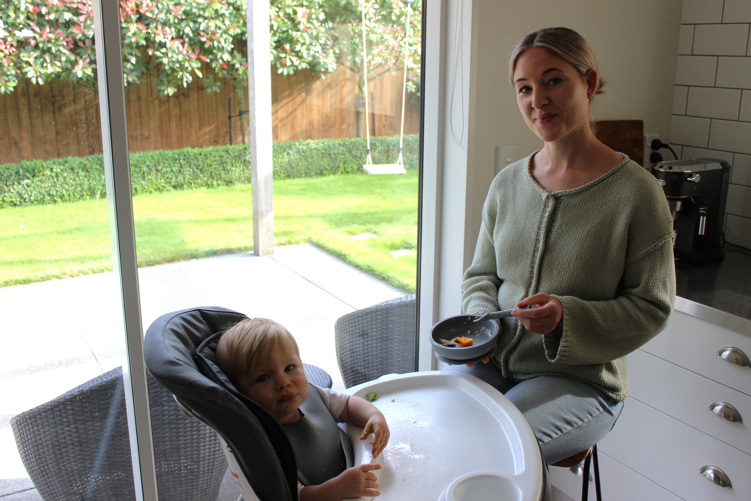 A woman with blonde hair and a beige sweater holding a bowl of food stands in a kitchen next to a child sitting in a high chair, inside a house with a large sliding glass door that opens to a backyard with green grass, bushes, and a wooden fence.