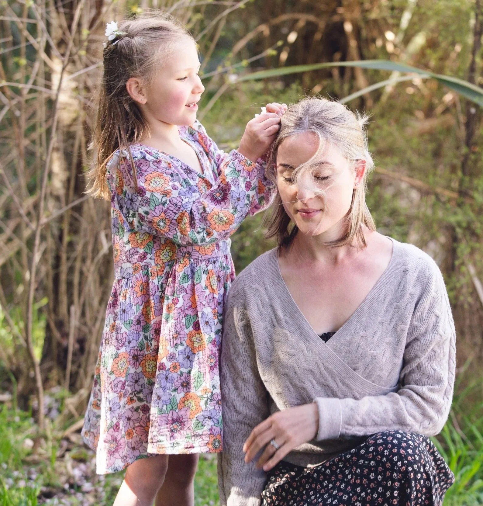A young girl with long hair and flowers in her hair, wearing a colorful floral dress, is playing a game with a woman who has blonde hair, wearing a light gray sweater, in an outdoor natural setting with trees and greenery in the background.