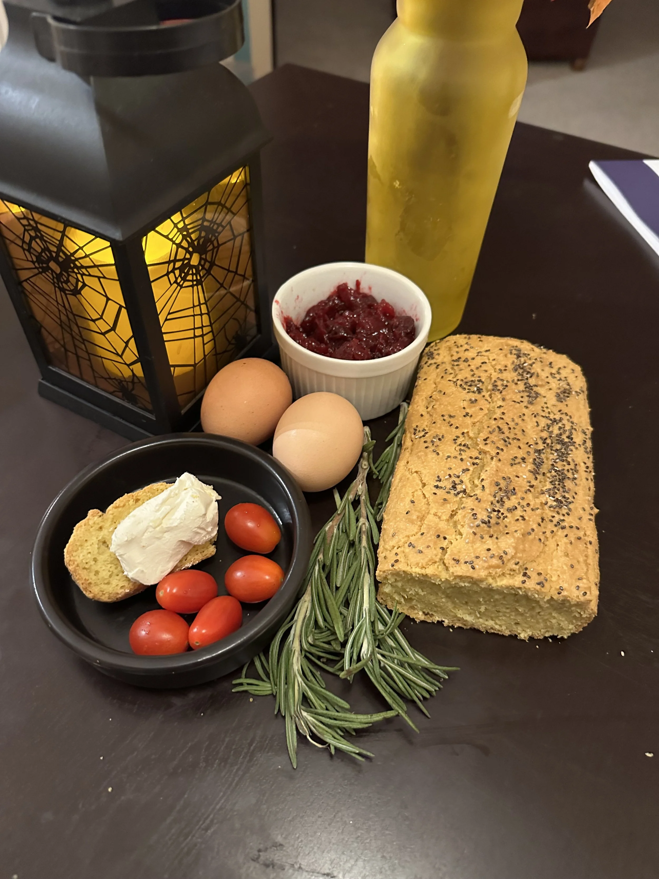 A breakfast spread including a loaf of bread topped with poppy seeds, a small bowl of cherry tomatoes with a slice of bread and cream cheese, two eggs, a jar of cranberry sauce, a sprig of rosemary, a yellow bottle, and a black lantern with a spiderweb design on the side.