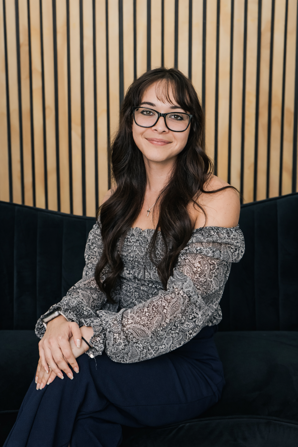 A woman with long dark hair, glasses, and a smile, sitting on a black velvet bench. She is wearing an off-shoulder patterned blouse and navy pants, with a cross necklace. The background features wooden vertical slats.