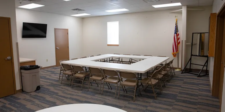 Empty conference room with tables arranged in a rectangle, beige chairs around the tables, a wall-mounted TV, an American flag, and a window with blinds.