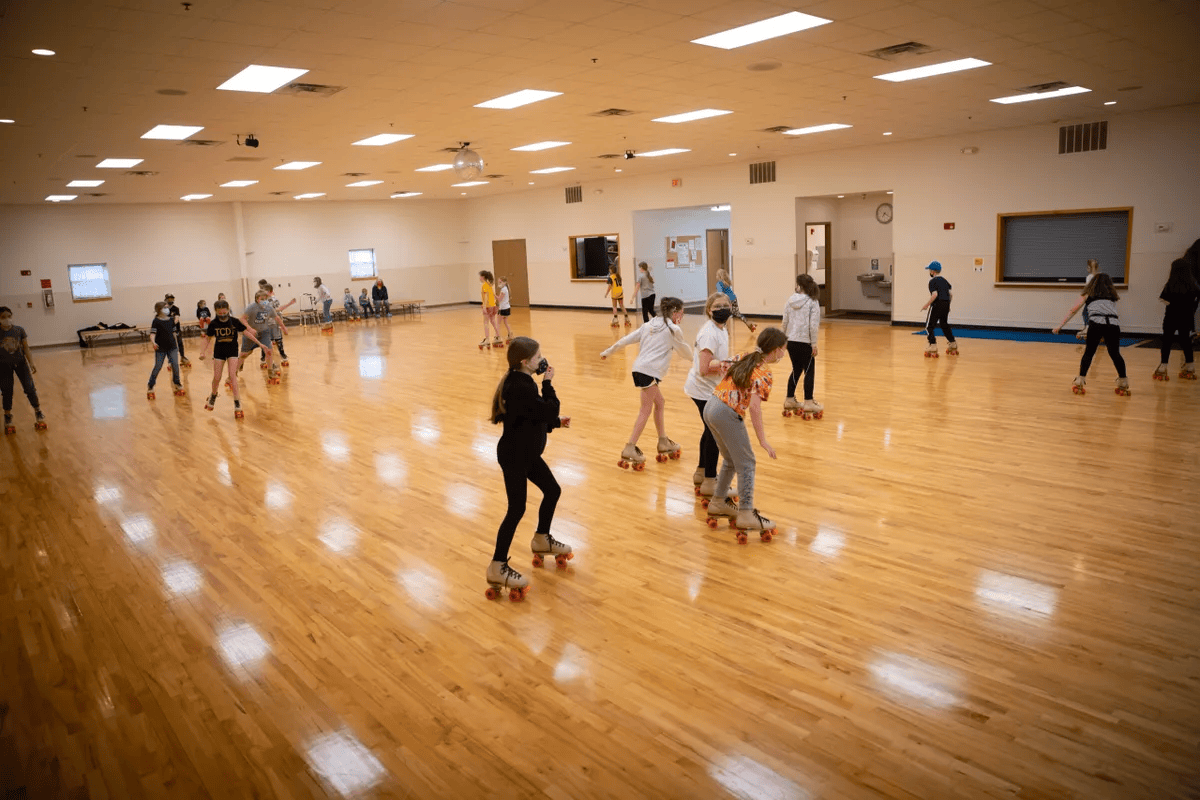 Children roller skating in a large indoor skating rink with wooden floors and white walls.
