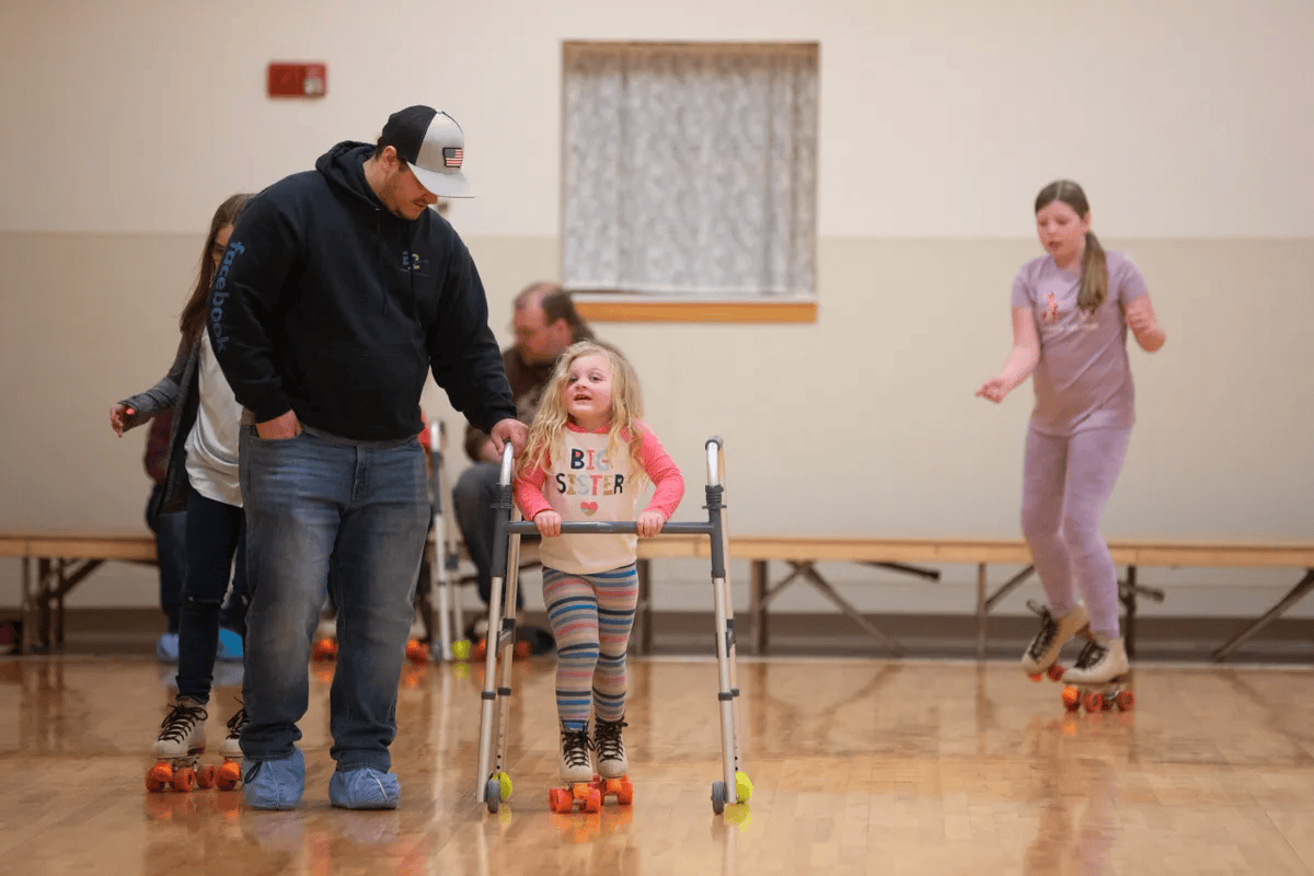A young girl using a walker is roller skating in an indoor rink, with an adult man helping her. Other children are roller skating in the background.