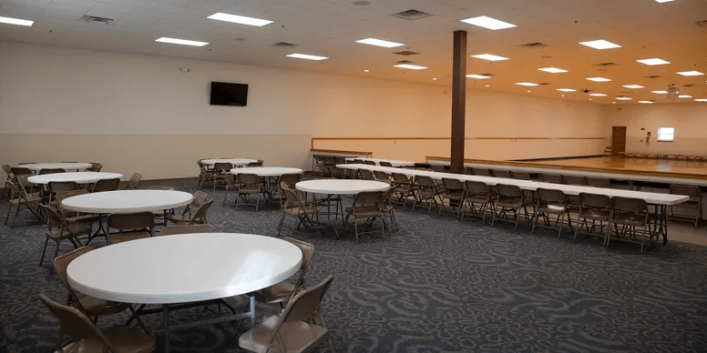 Empty banquet hall with round tables and chairs, carpeted floor, and a stage area in the background.