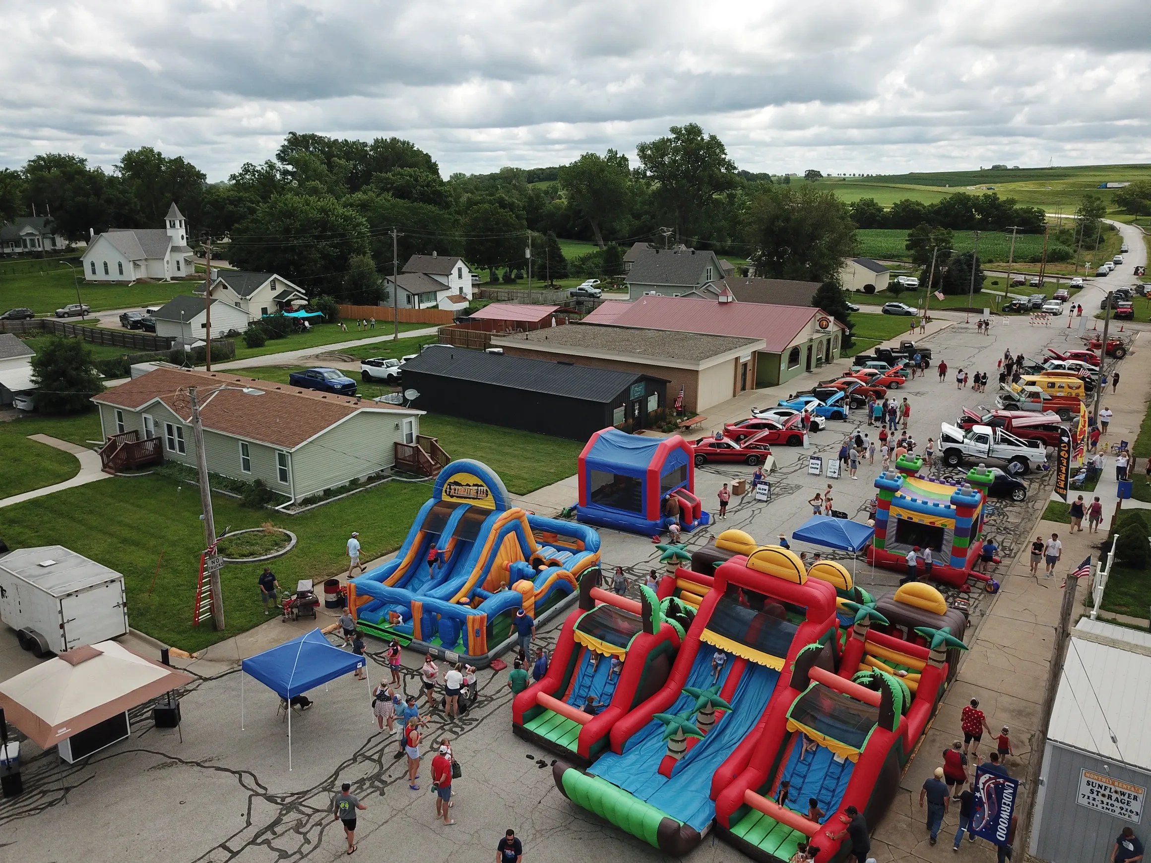 A street fair with multiple inflatable slides, people walking, and parked cars. Small buildings and houses line the street, with a cloudy sky overhead.