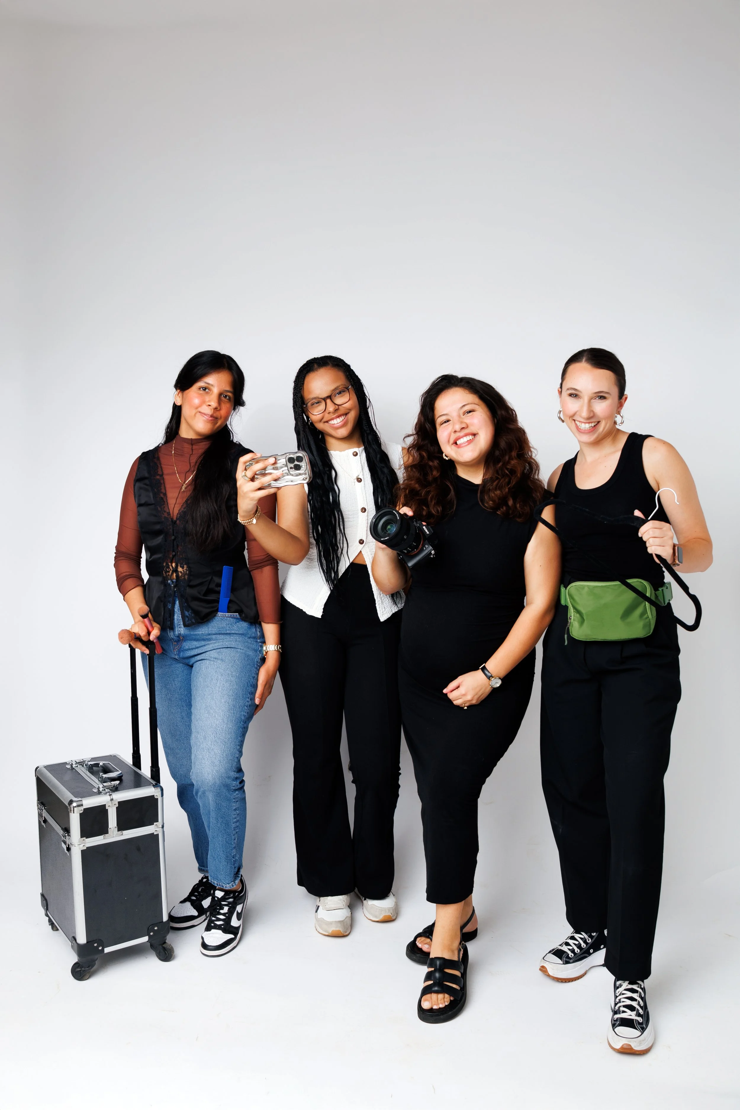 Four women smiling and holding cameras, phones, and a small rolling case standing against a plain white background.