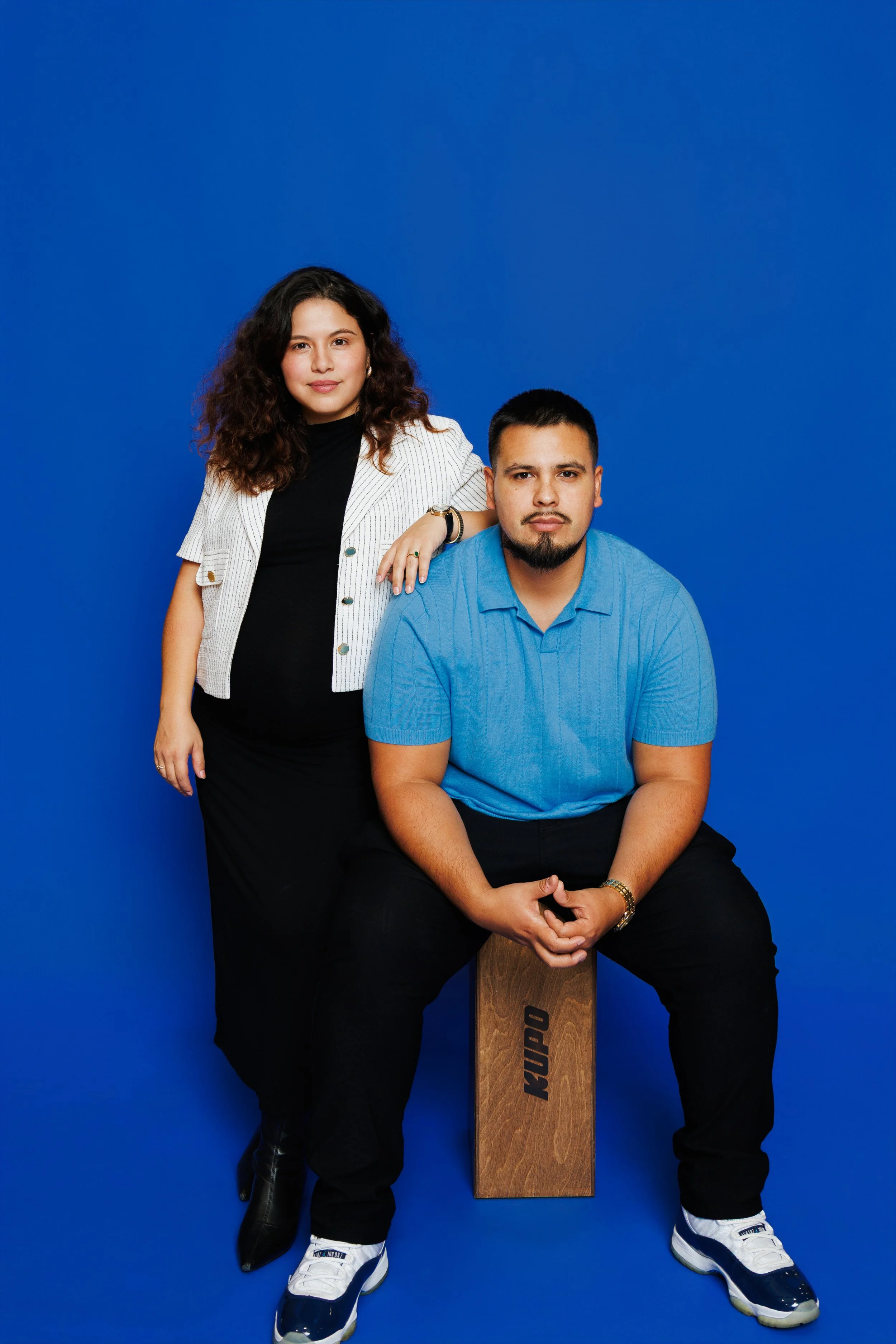A woman with curly hair wearing a black dress and white blazer standing next to a man with short hair, beard, wearing a blue polo shirt seated on a wooden box with the word 'KUPO' on it, against a bright blue background.