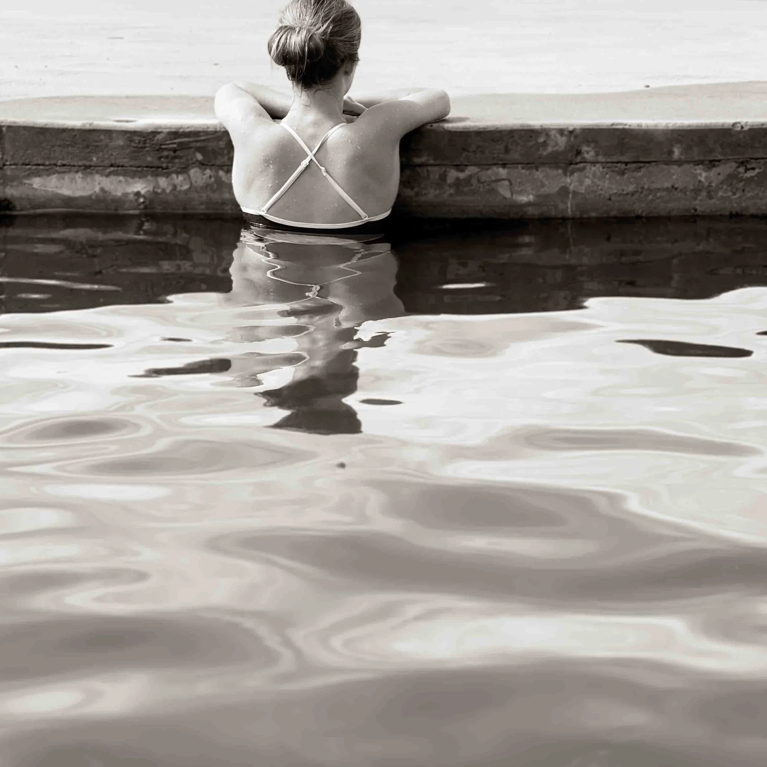 A woman leaning on a dock or pier, partially submerged in water, with her back to the camera.