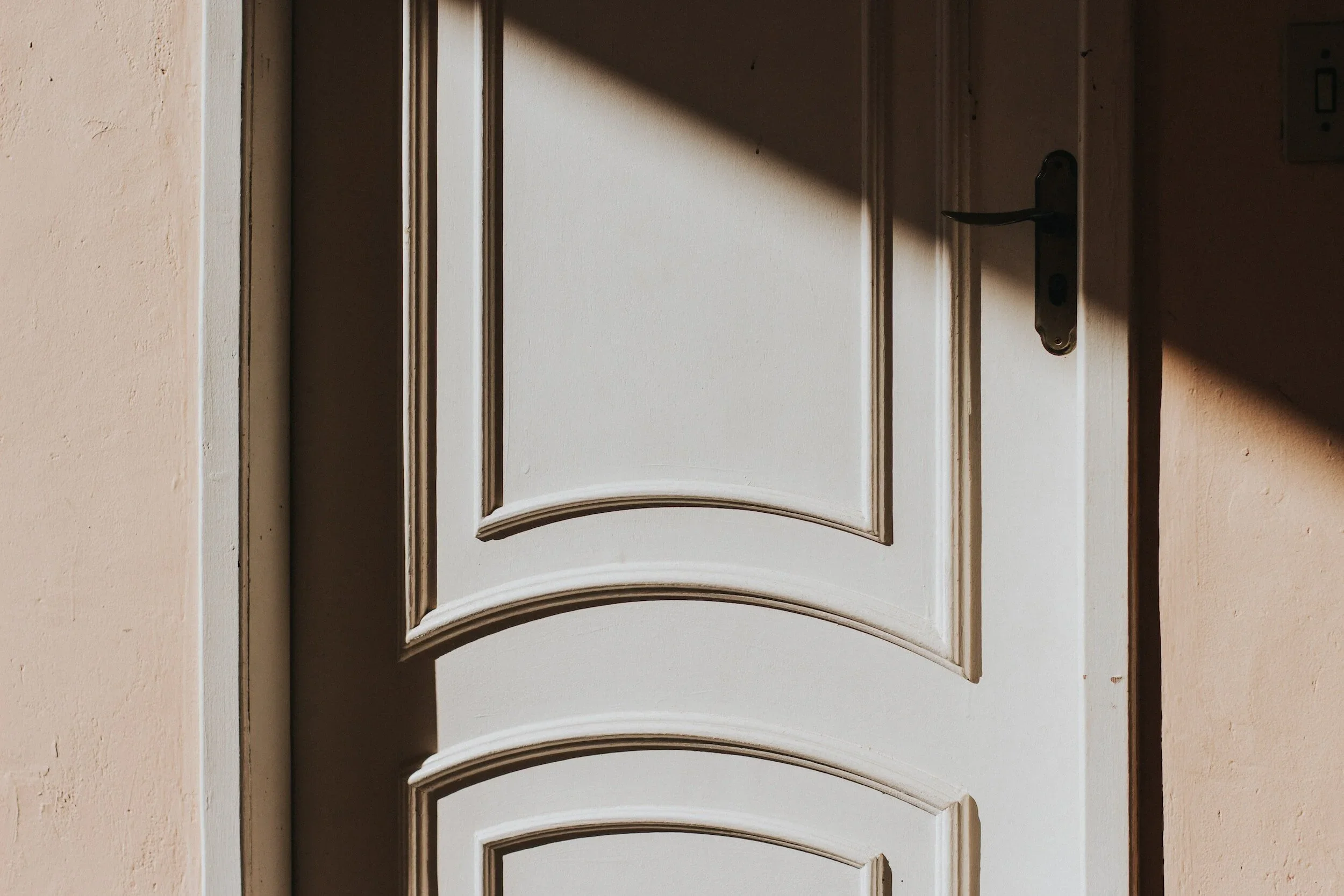 Close-up of a white door with a decorative panel design, partially closed, with a black door handle and lock, and a light switch on the side wall. Shadows are cast across the door.