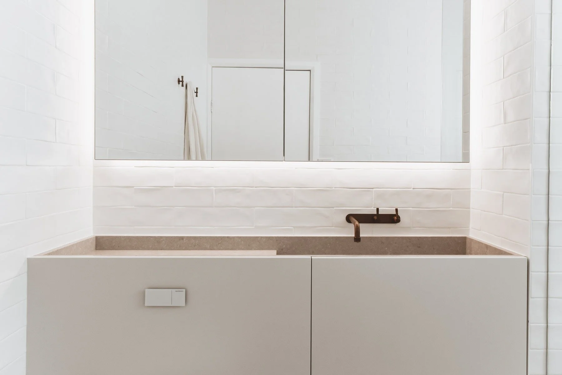 Minimalist bathroom with white brick walls, a large mirror, a floating vanity with a built-in sink, a copper faucet, and a mirror reflected in the background.