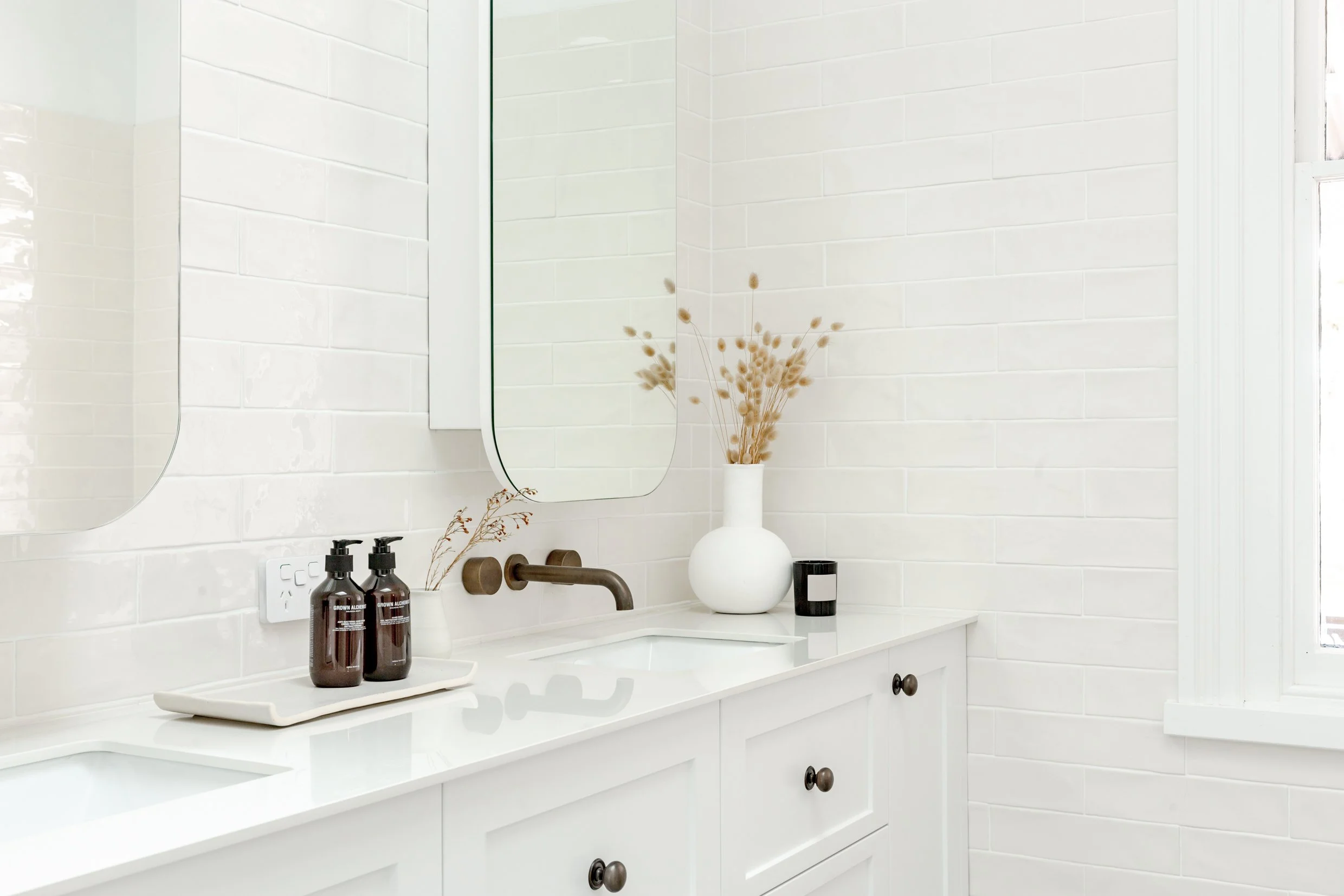A modern bathroom vanity with two sinks, a large mirror, decorative dried plants in a white vase, and two soap dispensers on a tray.
