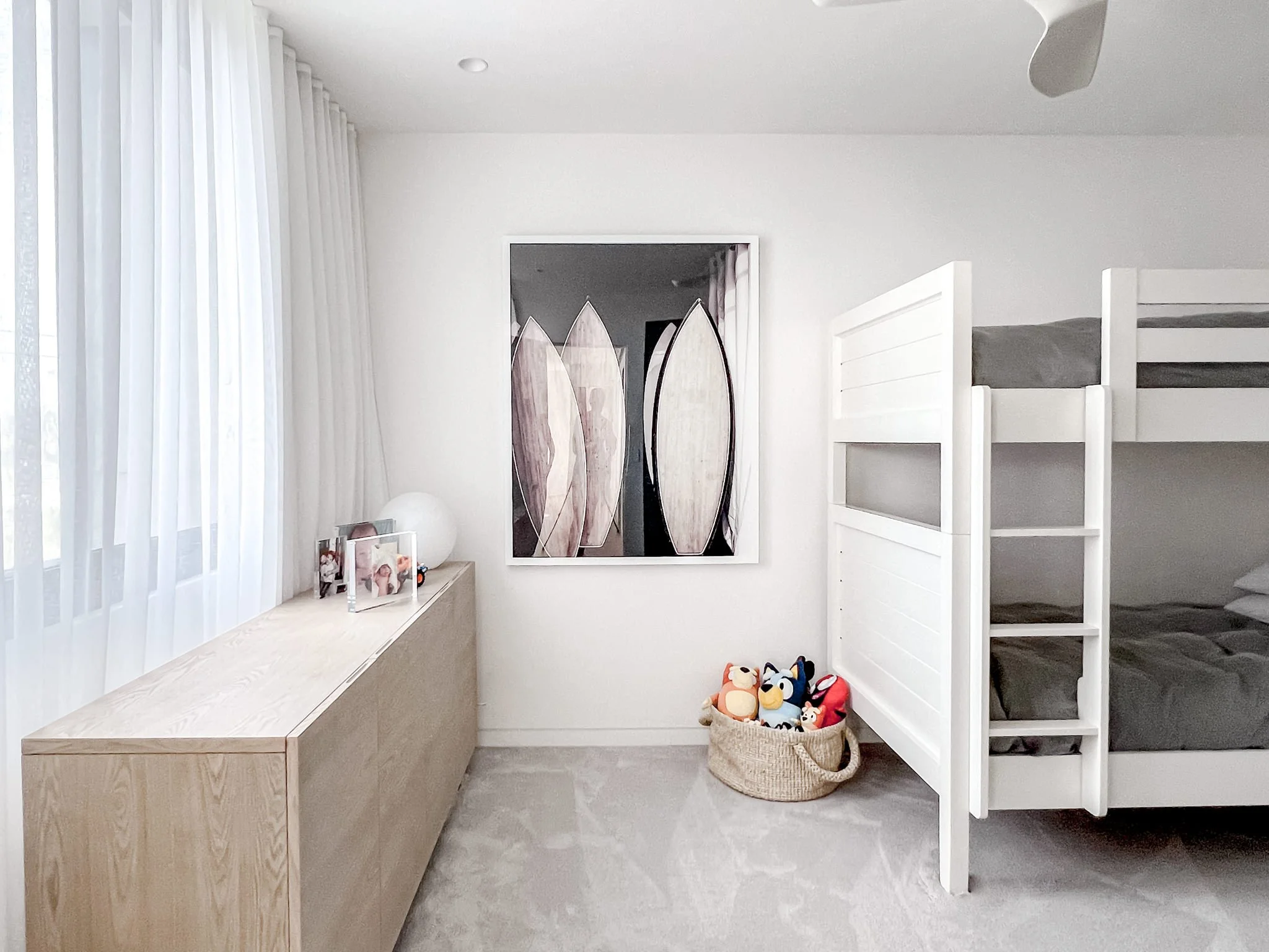A minimalist children's bedroom with white walls, gray plush carpet, and white furniture. There is a bunk bed with gray bedding, a light wood sideboard with framed photographs and a round lamp, a colorful plush toys basket, and a large framed abstrac