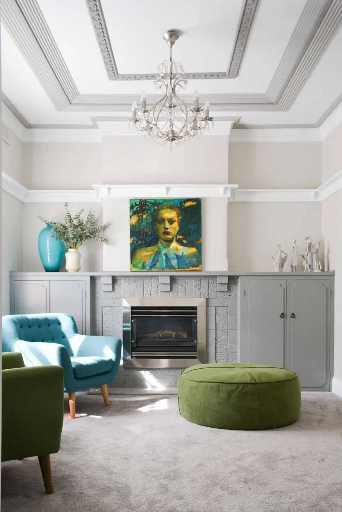 Gather Studio - Bar Beach Project. Living room with a fireplace, gray cabinetry, a colorful portrait above the fireplace, a blue armchair, a green ottoman, and decorative items including a vase with greenery and art figurines.