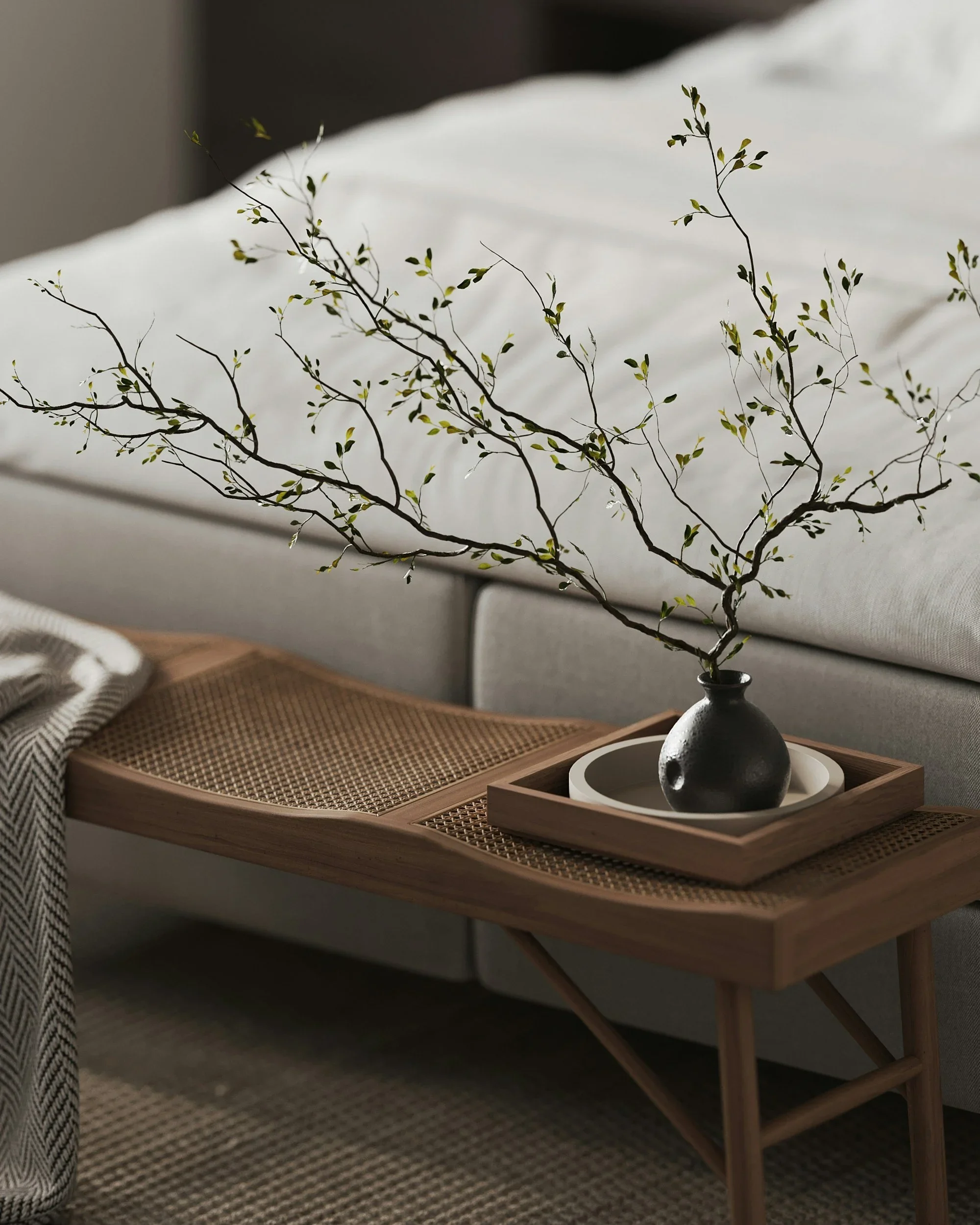 Decorative branch with small leaves in a black vase on a wooden tray, placed on a wooden table next to a beige sofa.