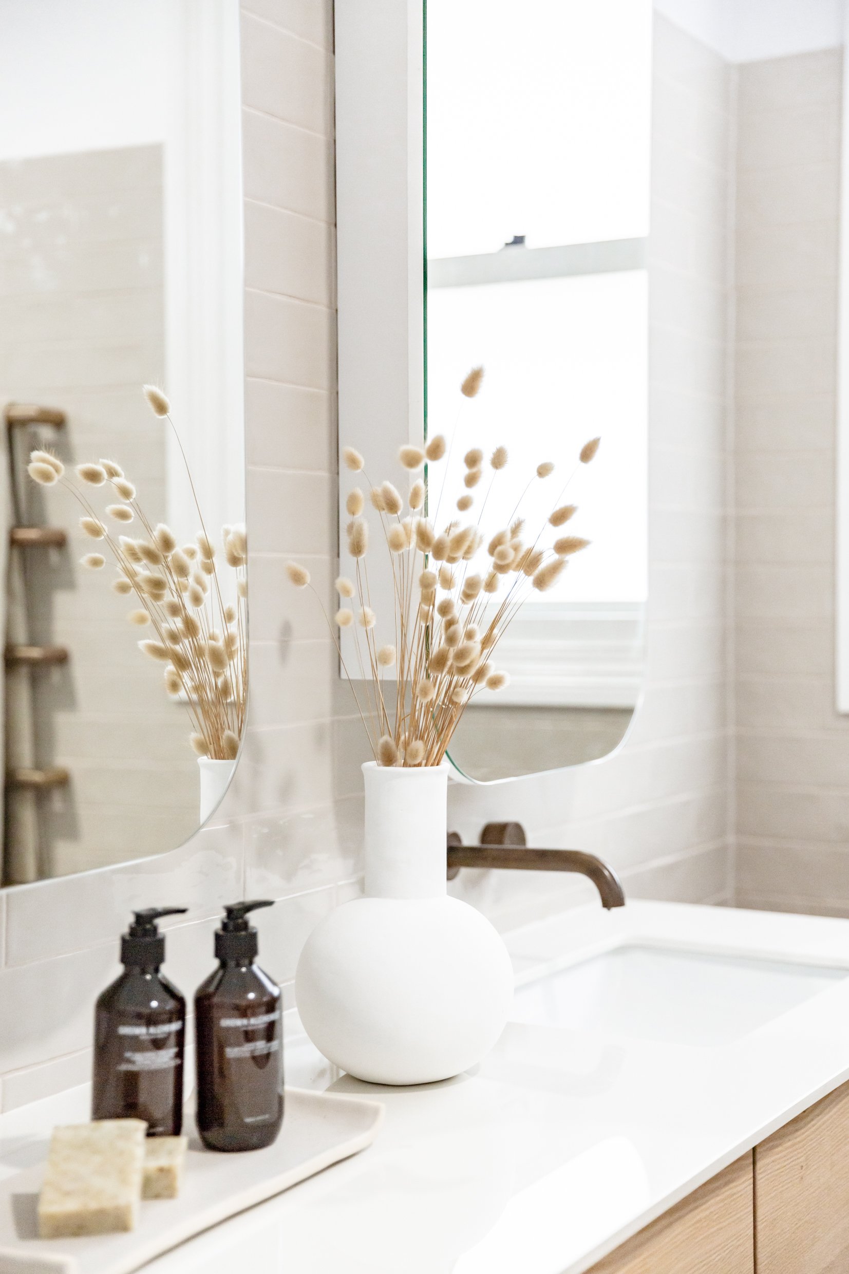 Gather Studio - Urban Farmhouse. Bathroom vanity with a white vase containing dried flowers, two brown soap dispensers, and a soap bar on a tray, reflected in a mirror.