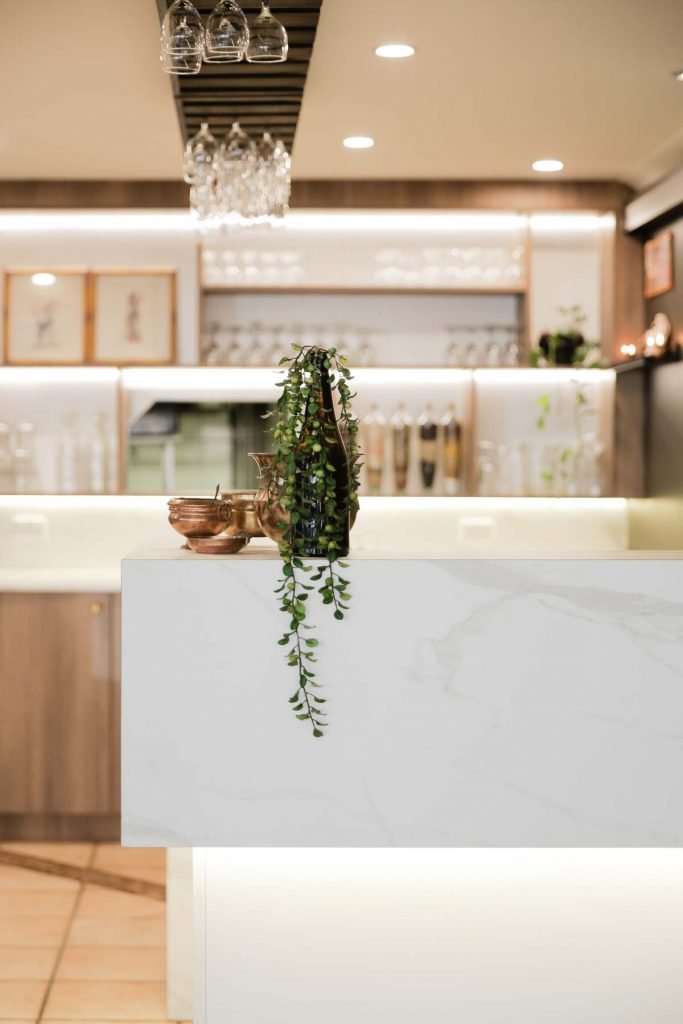 Gather Studio The Only Place Indian Restaurant.Interior of a modern restaurant or cafe with a white marble counter, green hanging plant, and copper bowls. Glassware is hanging upside down from the ceiling, and soft lighting creates a cozy atmosphere.