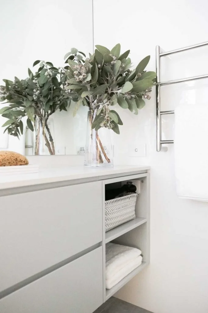 Gather Studio - Ensuite Renovation. A white dresser with two shelves holding a basket and folded towels, topped with two glass vases containing green leafy branches.