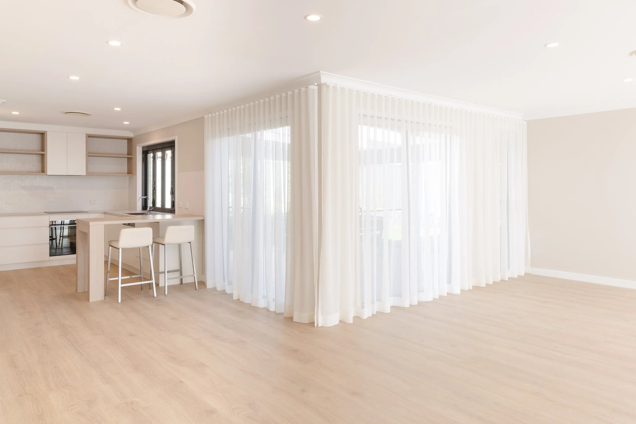 Empty living room with light wood flooring, white walls, large window with sheer white curtains, and open kitchen with white cabinetry, a small dining island, and two white chairs.