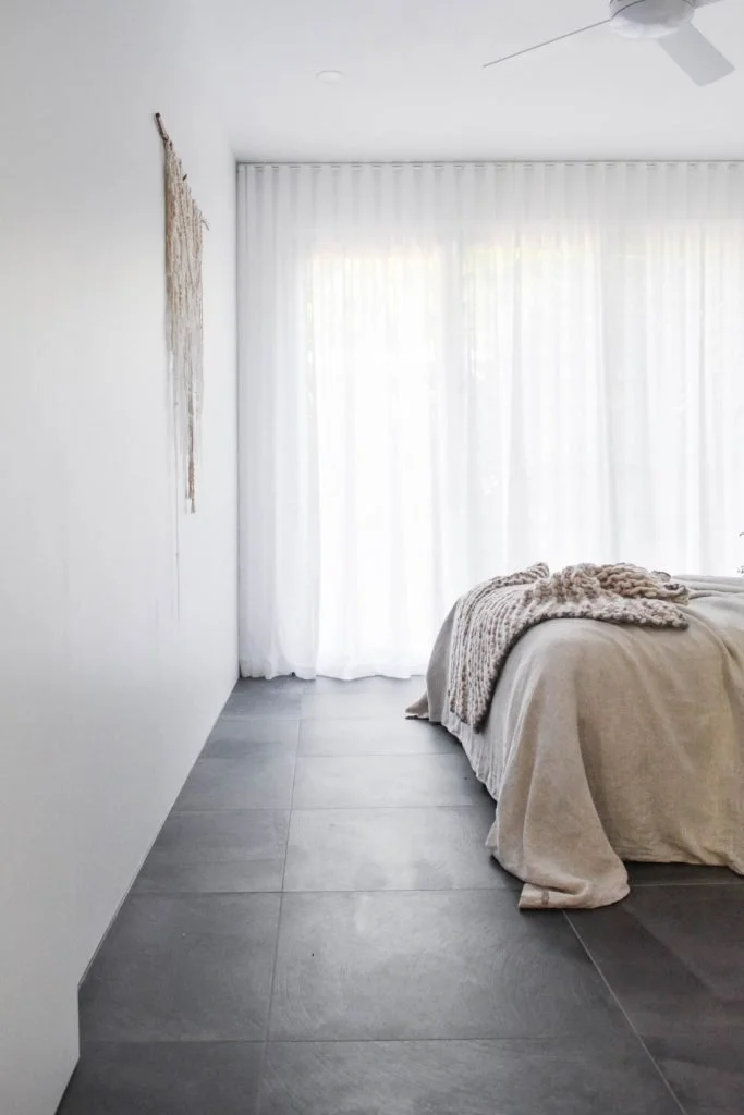 Gather Studio - Garage Master Suite Conversion. A bedroom with white walls and large gray tiled floor, beige bed with a textured blanket, and white curtains covering a window or sliding doors.