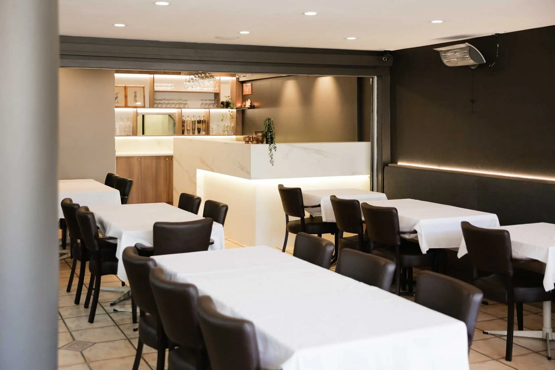 Empty restaurant with tables covered in white tablecloths and black chairs, modern decor with a marble counter and warm lighting.