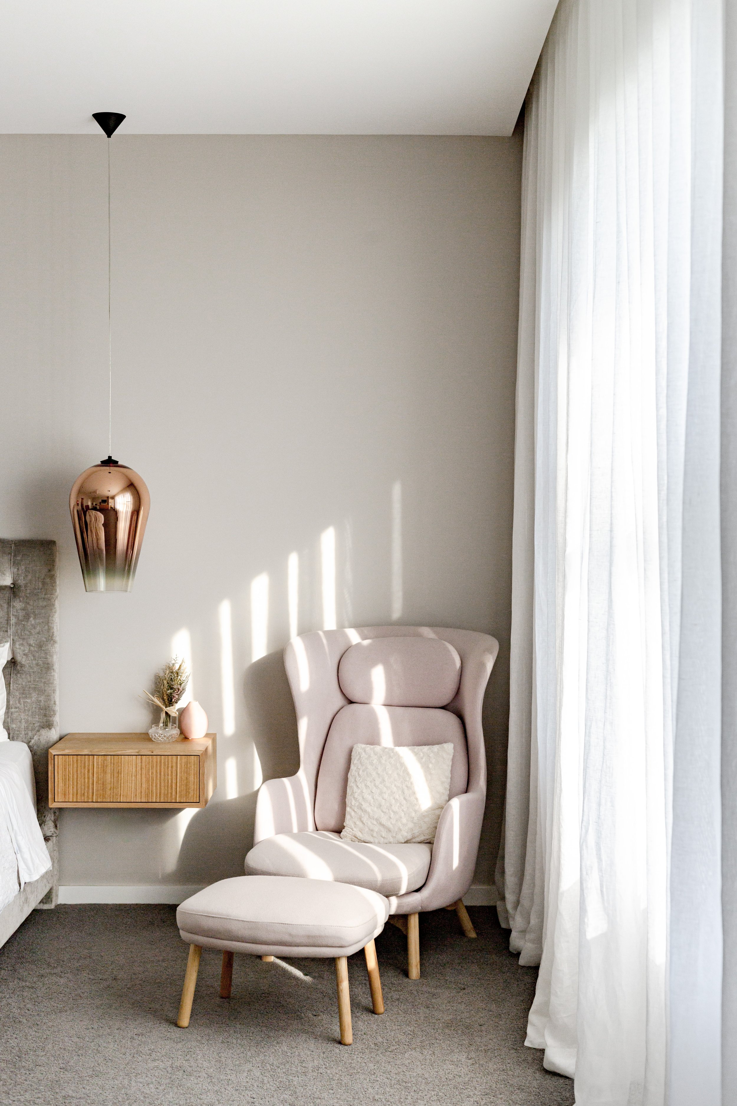 A cozy corner in a bedroom with a high-backed armchair and matching footrest, a floating wooden nightstand with a small decorative item, and white curtains letting in natural light. A pink pendant light hangs from the ceiling.