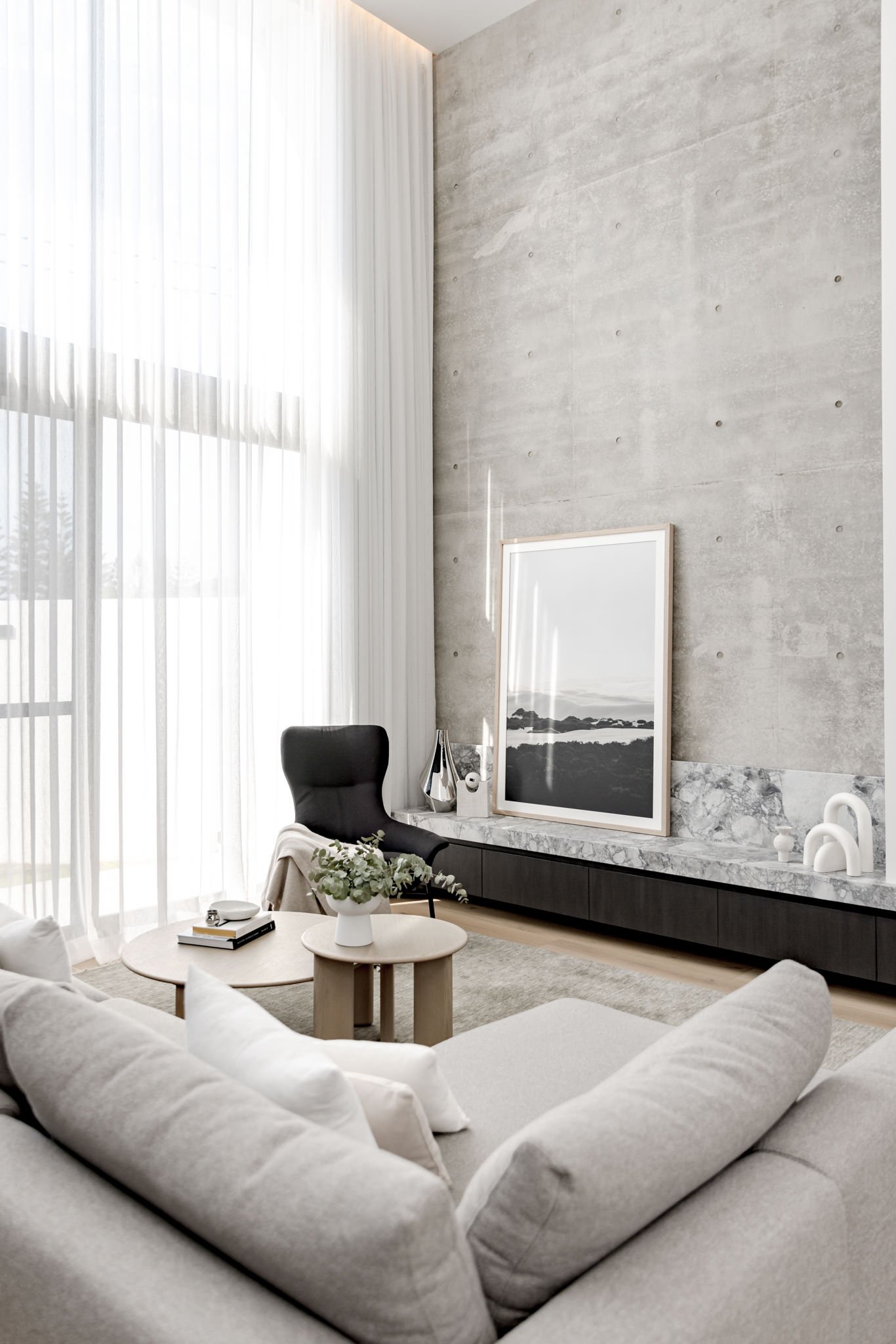 Modern living room with large window and sheer curtains, beige sofa with pillows, small round coffee table with flowers, black chair, and a built-in marble shelf with decorative items and a framed black-and-white landscape photo.