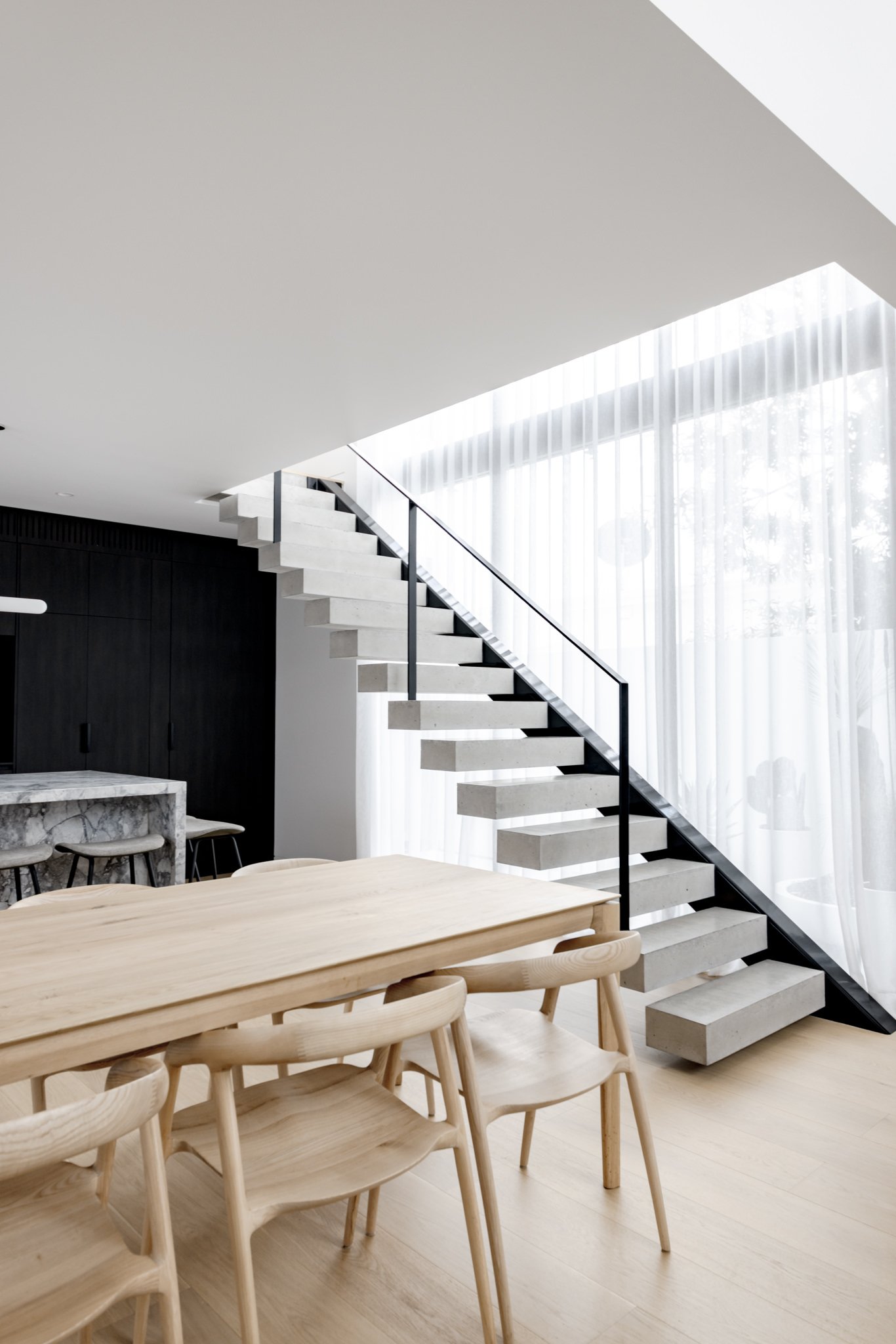 Modern dining area with a wooden table and chairs, black staircase with concrete steps and metal railing, large window with sheer curtains, and minimalist design.