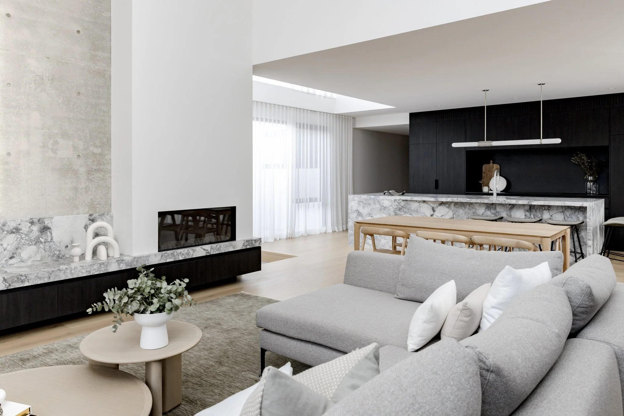 Modern living room with grey sectional sofa, wooden coffee tables, and a fireplace with marble surround. Open kitchen with marble island, black accent wall, and dining area with wooden table. Large windows with sheer white curtains.