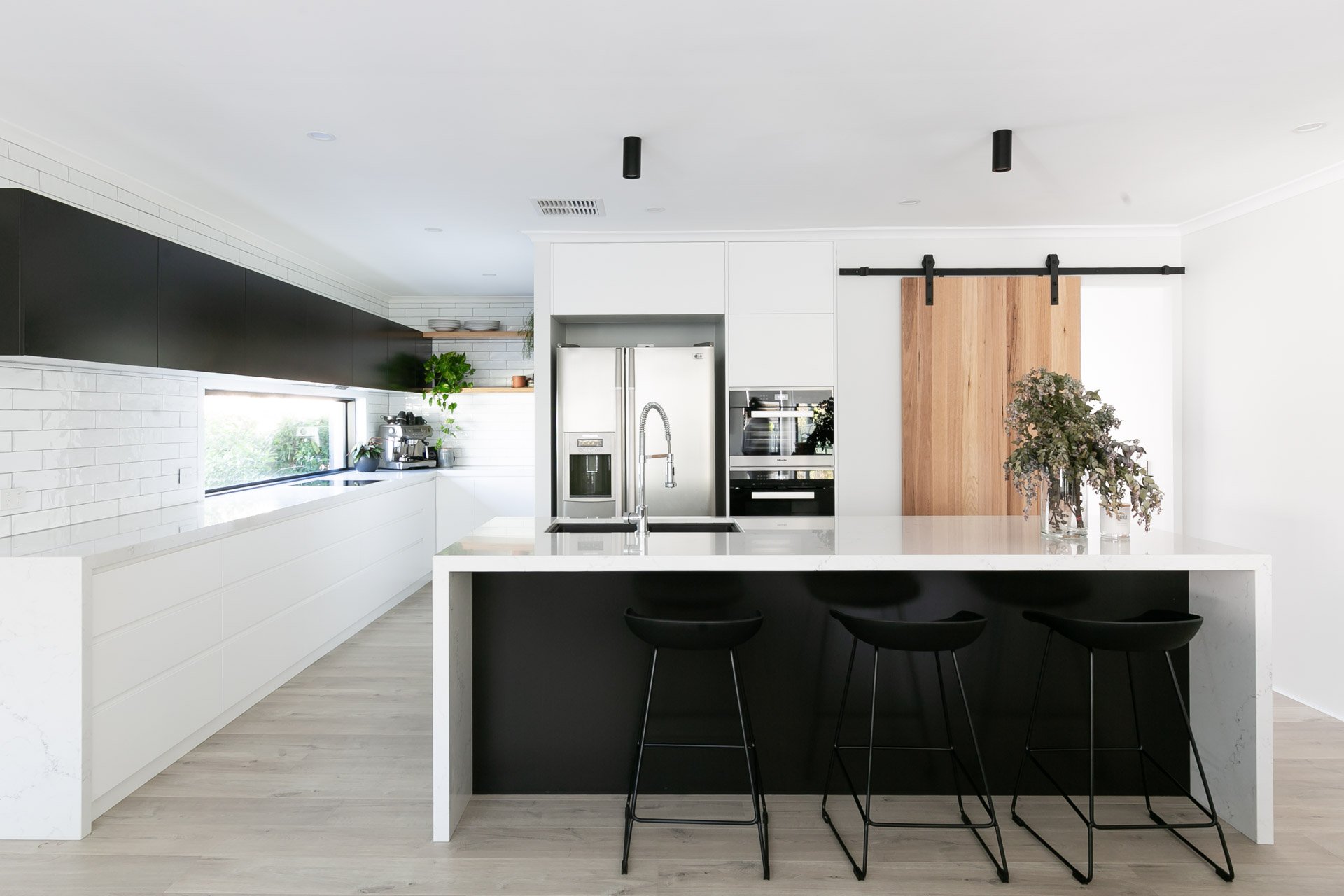 Gather Studio - Minmi Road Residence. Modern kitchen with white countertops, black and white cabinetry, stainless steel appliances, a black sliding barn door, and a large island with bar stools.