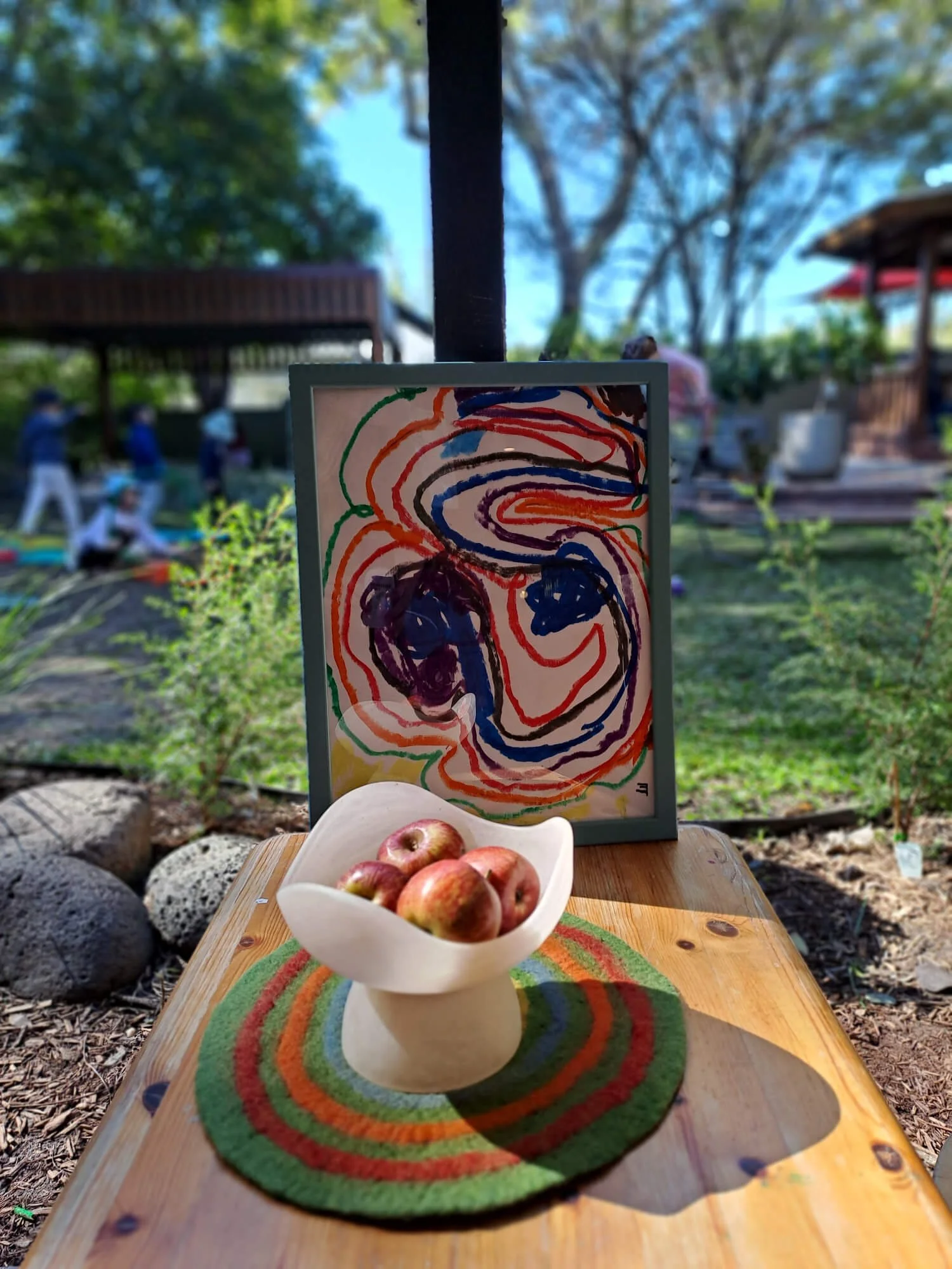 A wooden table outdoors holding a bowl of apples, a circular multicolored felt mat, and an abstract framed artwork with swirling lines and bold colors, with a garden and people walking in the background.