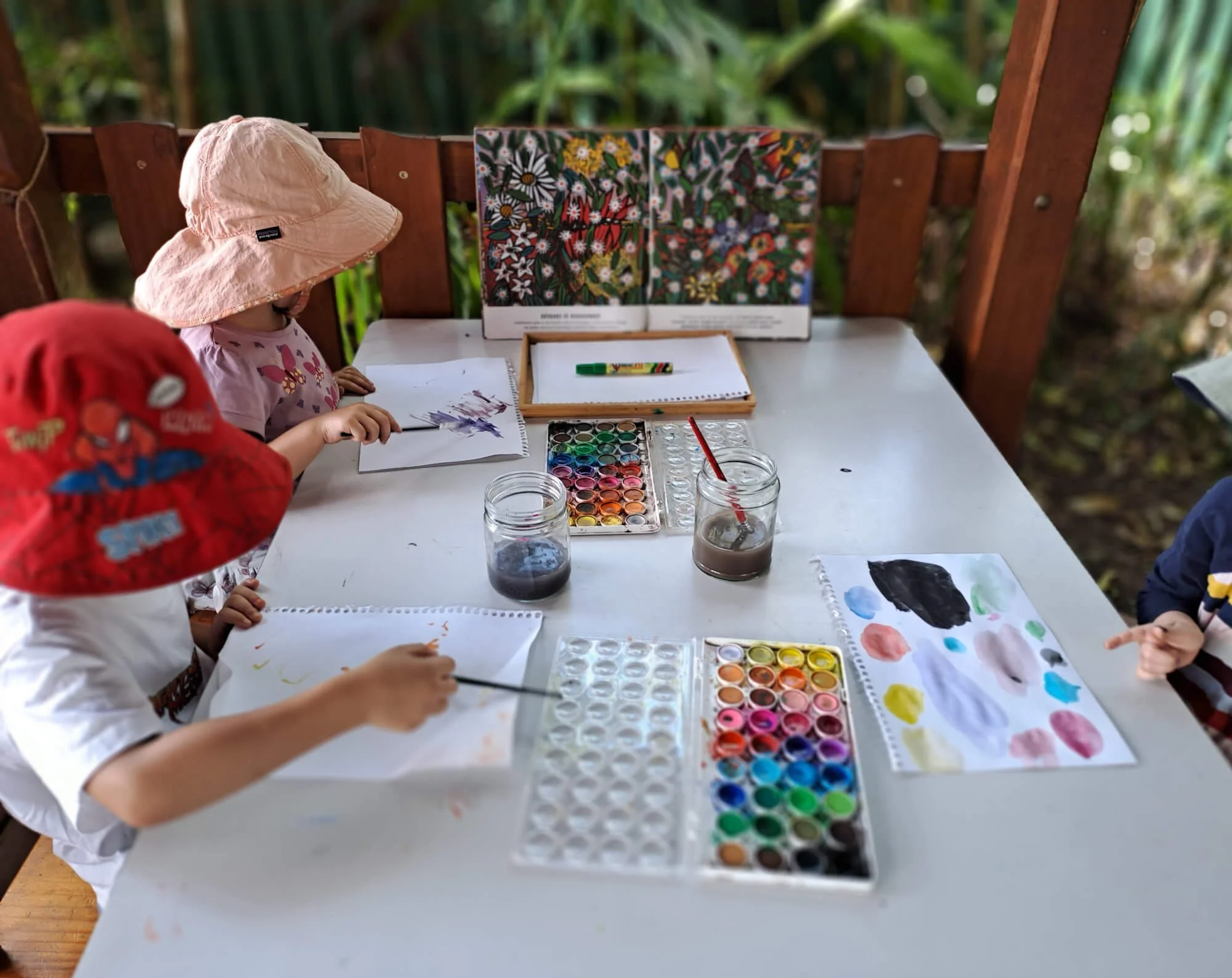 Children at Ballymore Kindy, Herston, painting and drawing at a white table outdoors with watercolour paints, brushes, and drawings, wearing hats to protect from the sun.