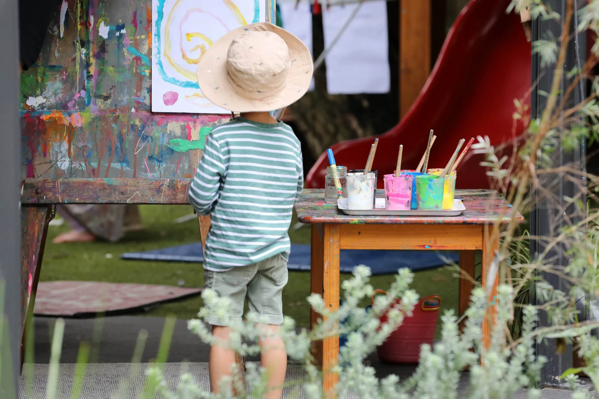 A young child is playing outdoors at Ballymore Kindy, Herston is wearing a straw hat and a green and white striped shirt, is facing a colourful abstract painting on an easel.