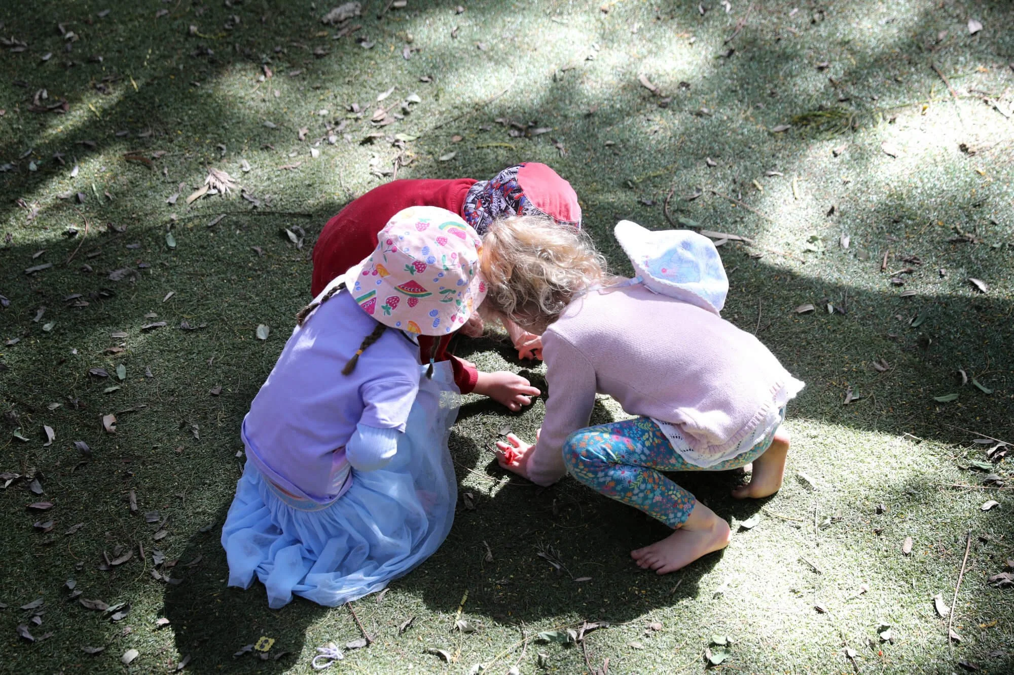 Three children crouching on grass, examining or playing with something on the ground outdoors.