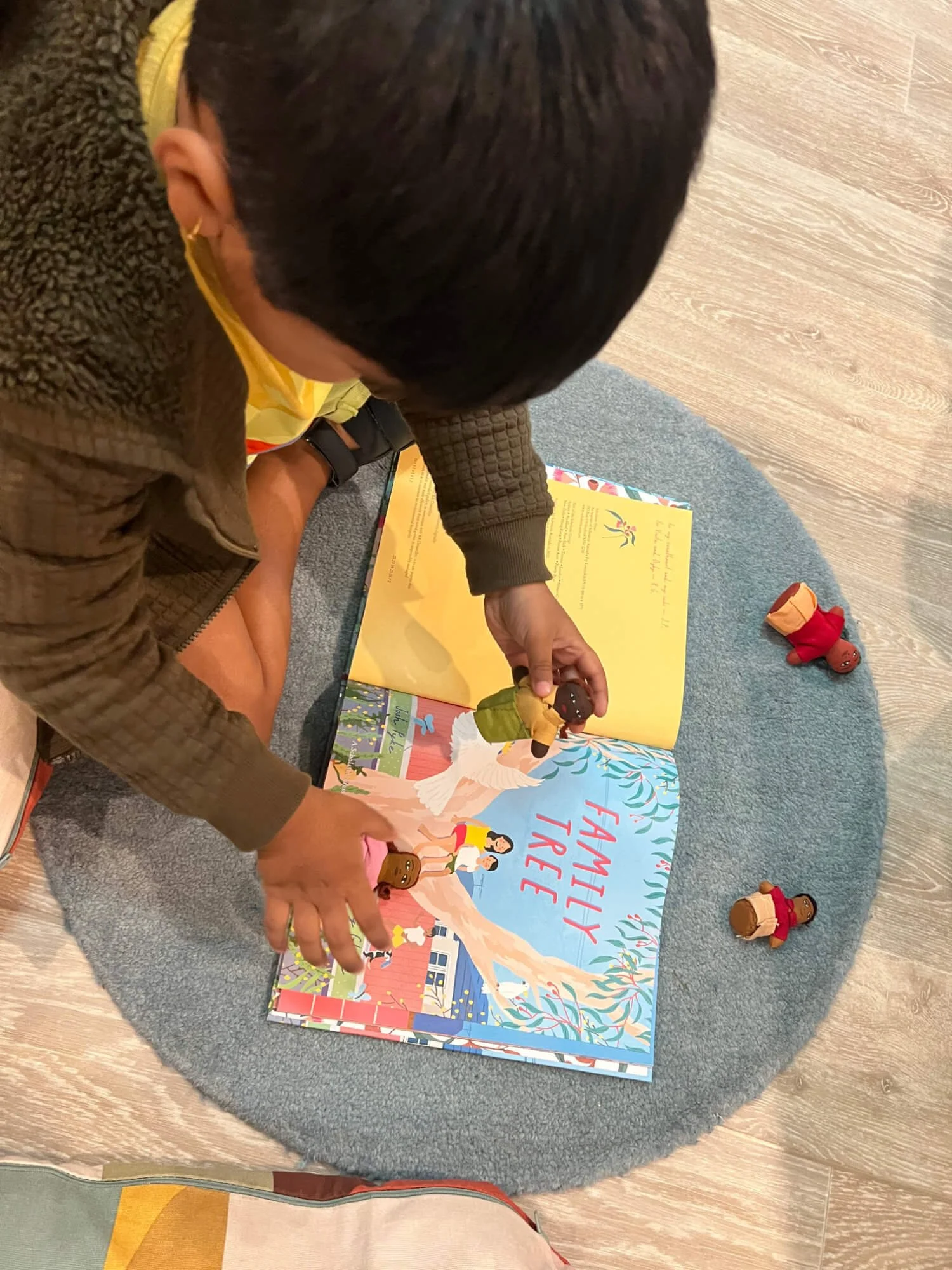 A child sitting on a gray round rug at Ballymore Kindy, Herston, reading a children's book titled 'Family Tree' with paper figurines representing family members placed on the rug.