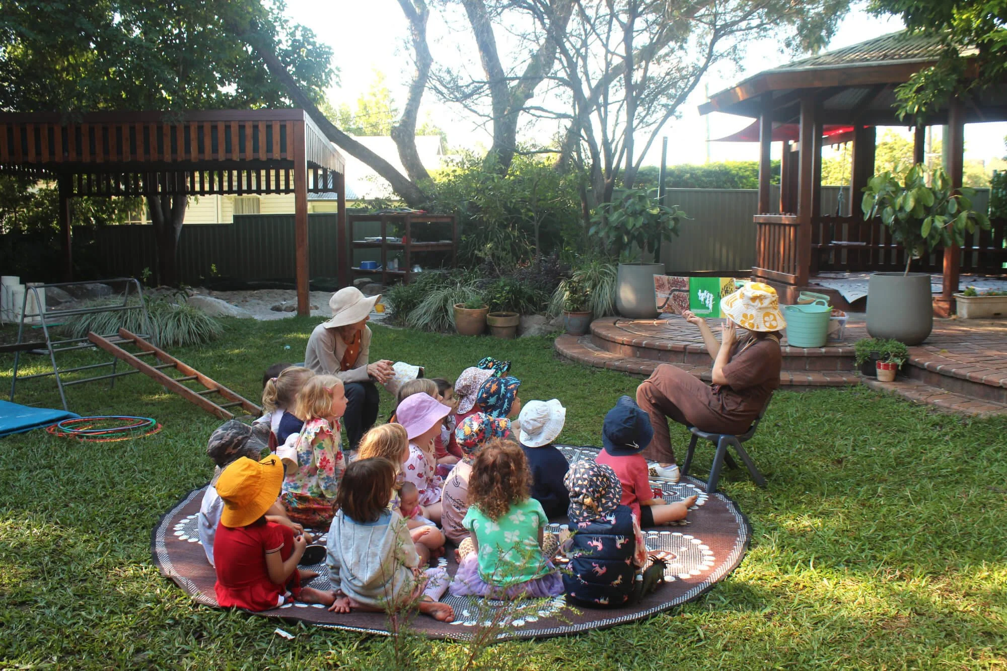 A woman reading a book to a group of children sitting on a large outdoor rug in a backyard with trees, plants, and a wooden gazebo.