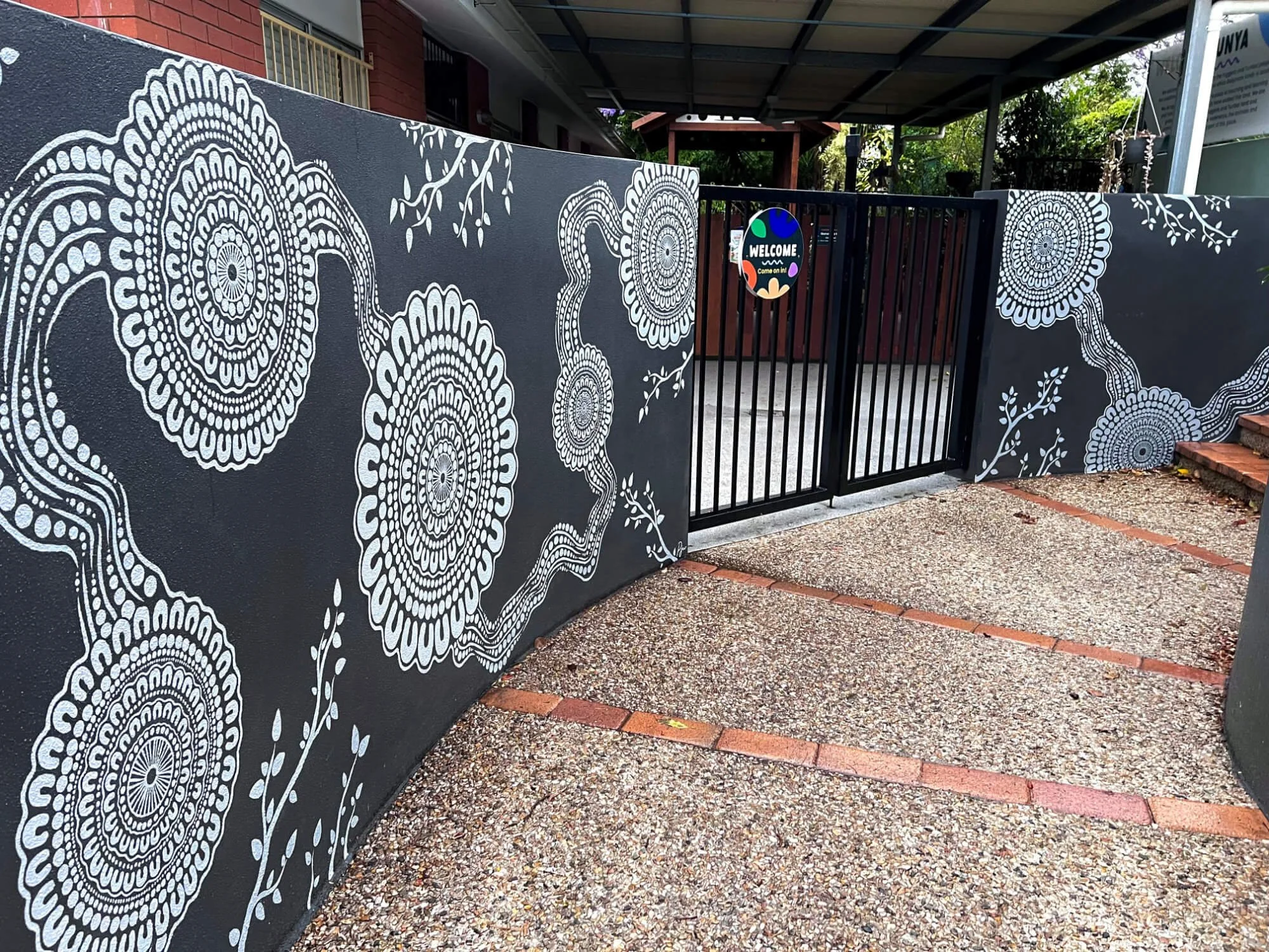 A black gate with a colorful welcome sign, flanked by black walls with white floral murals, leading to an outdoor area with trees and a brick walkway.