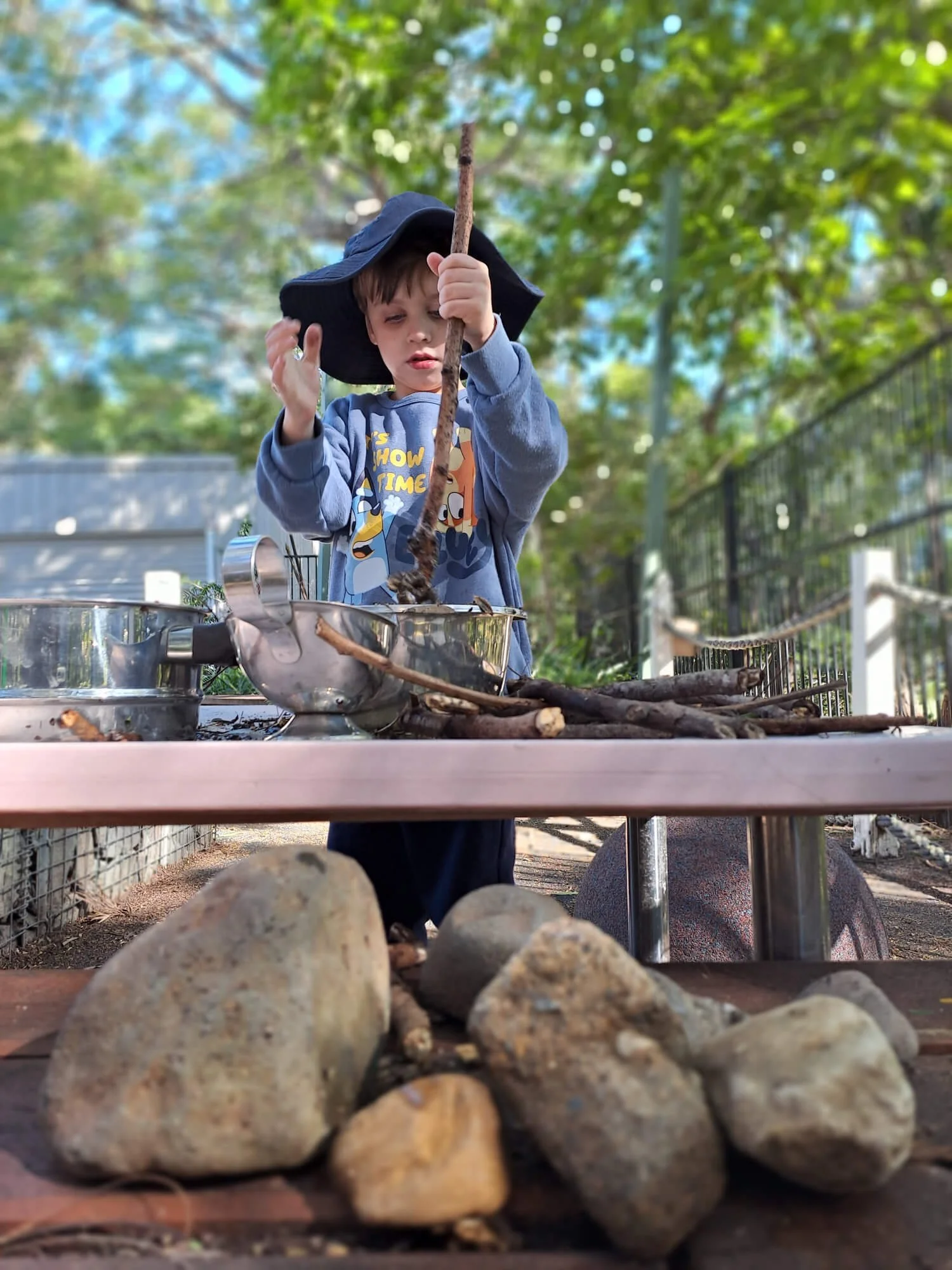 A young boy wearing a blue sweatshirt and a wide-brimmed hat, standing at an outdoor table, looking at a stick he is holding, with a background of green trees and bright sky.