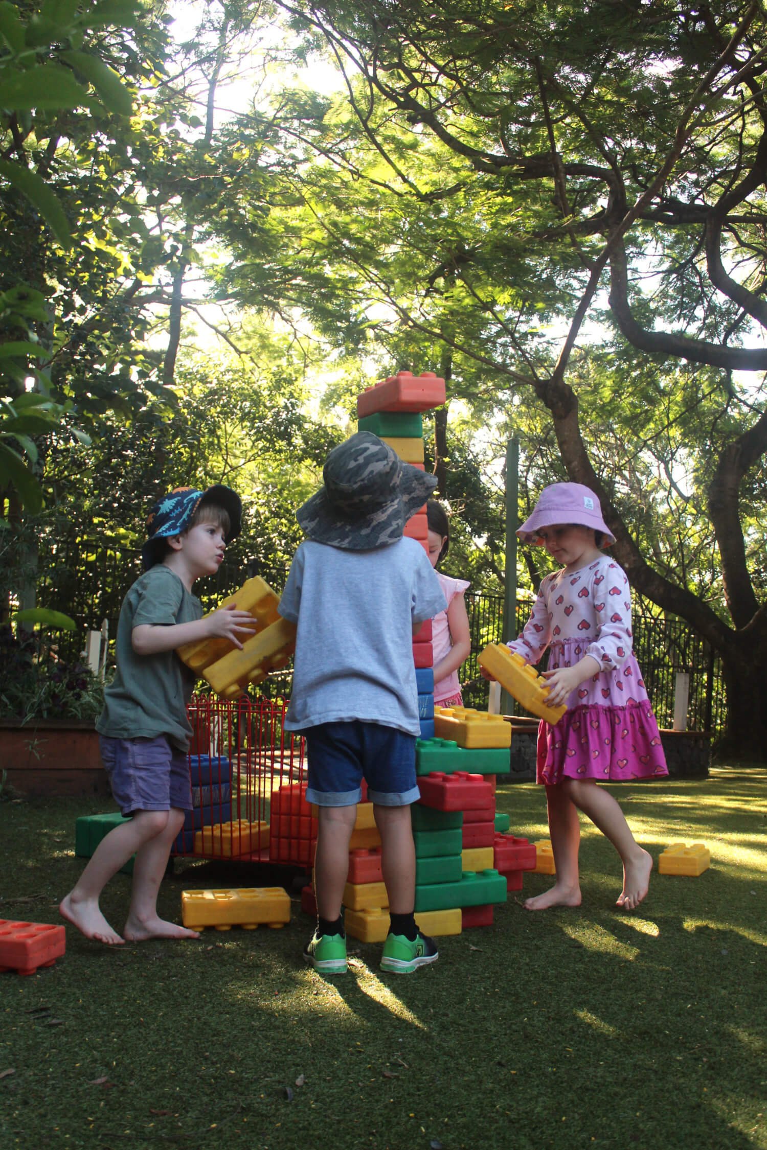 Children playing with large colorful building blocks outdoors in a grassy area surrounded by trees.