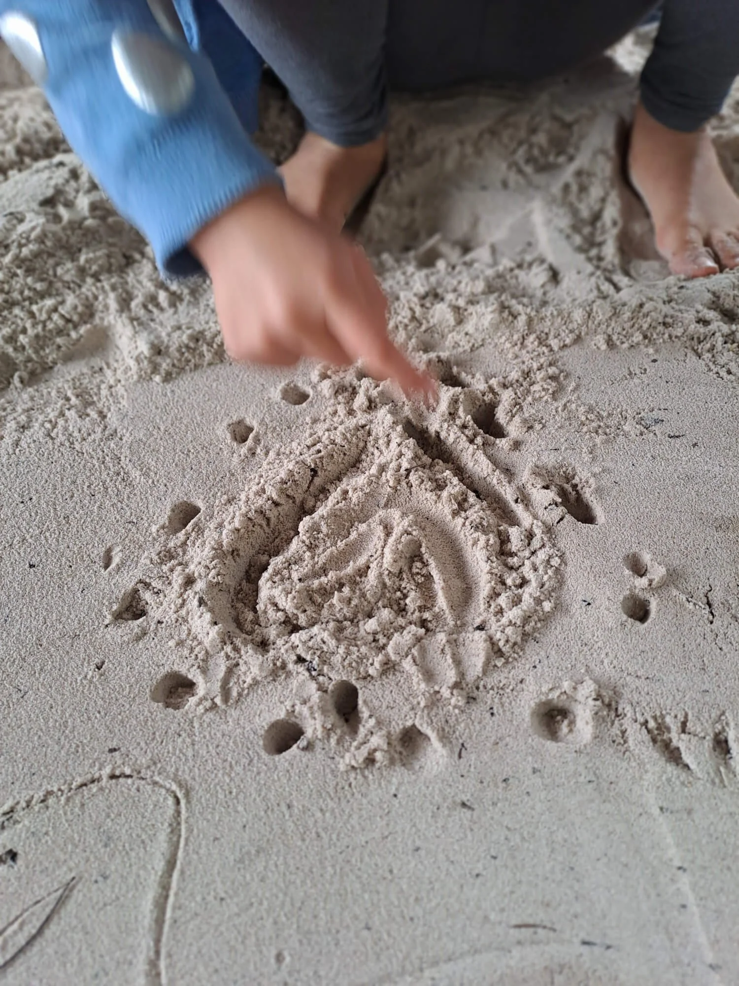 Child drawing a picture of a sailboat in the sand with a finger, surrounded by small hole marks in the sand.